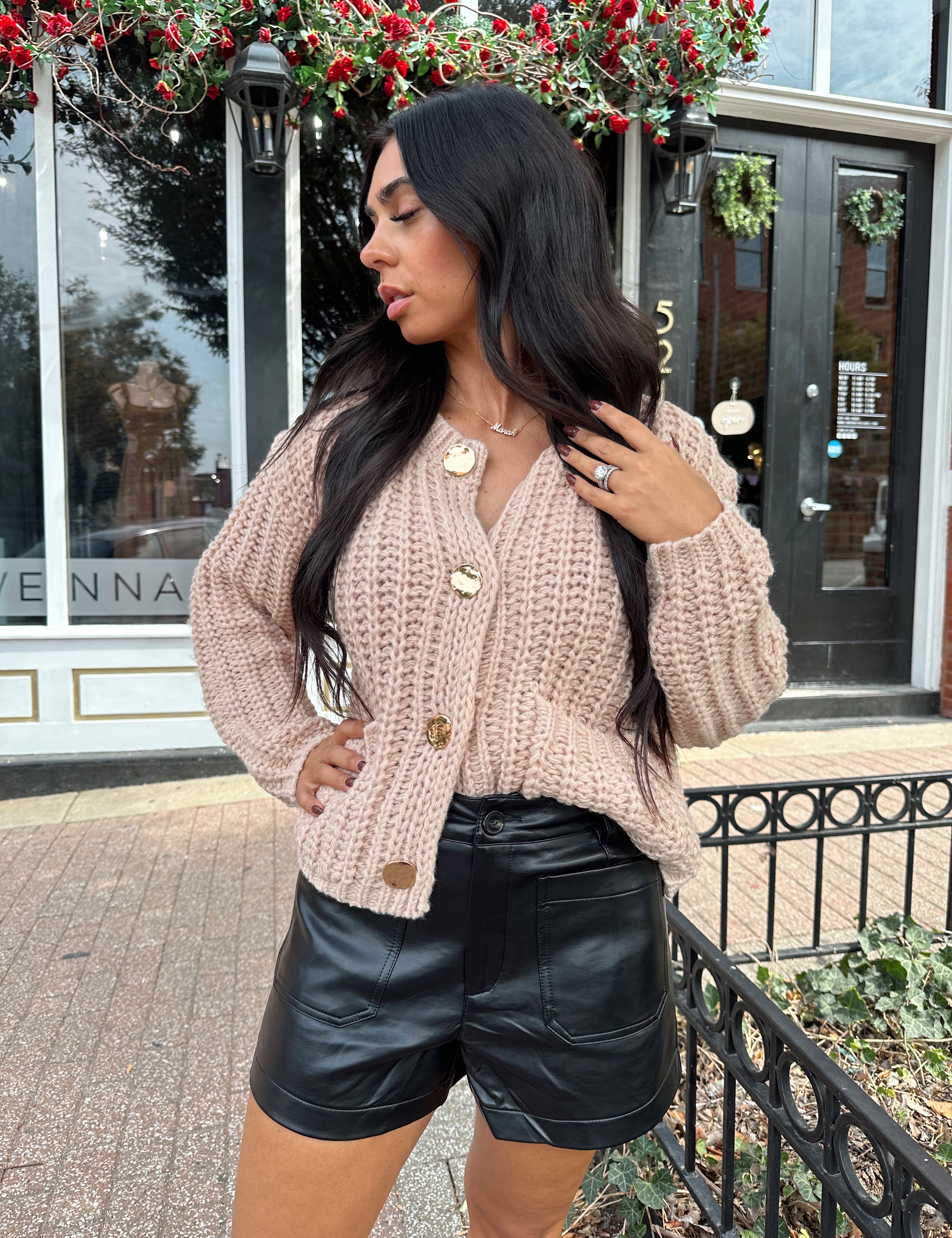 Woman wearing oatmeal colored knit chunky cardigan with gold button closure and functional pockets.