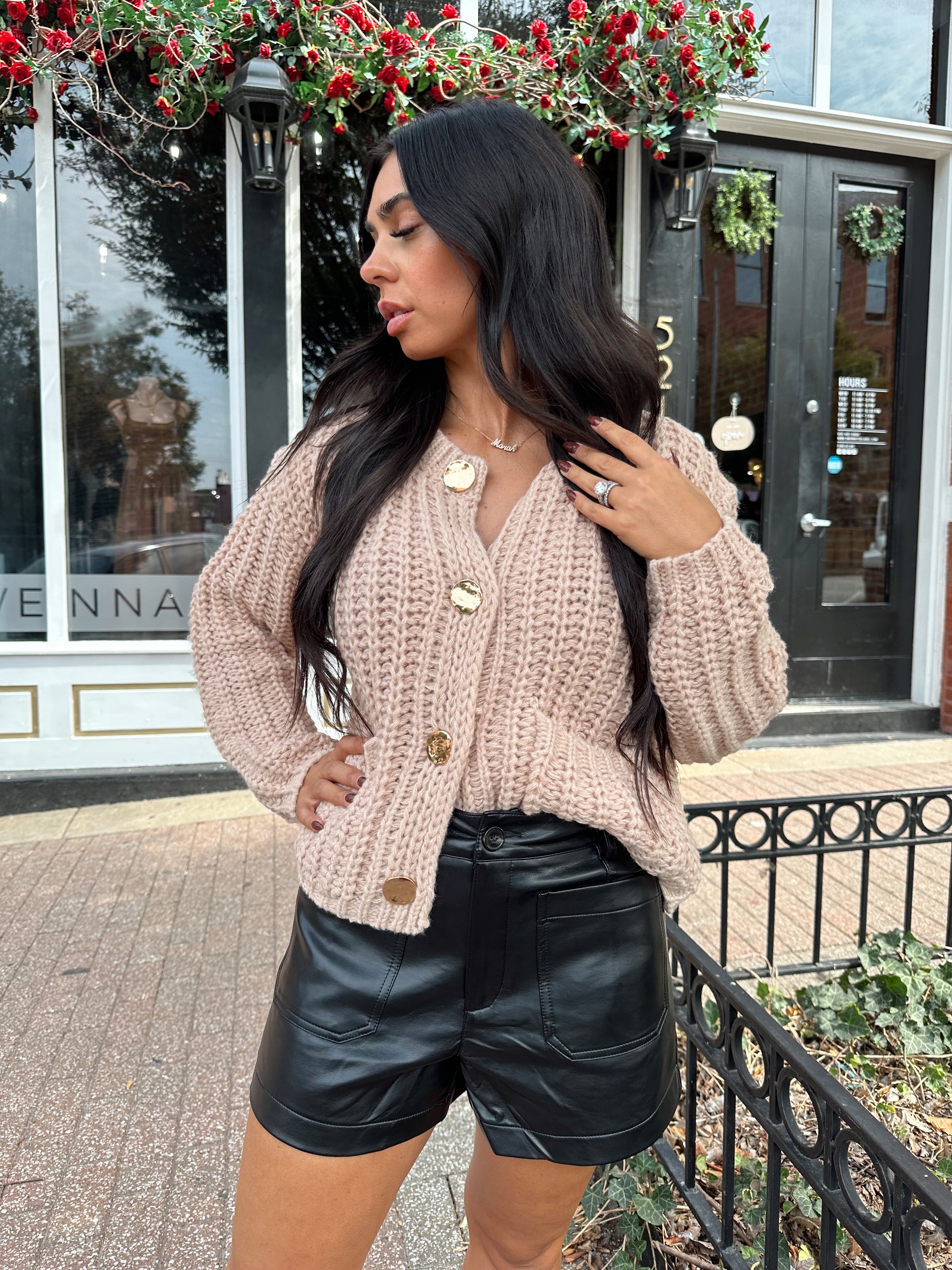Woman wearing oatmeal colored knit chunky cardigan with gold button closure and functional pockets.
