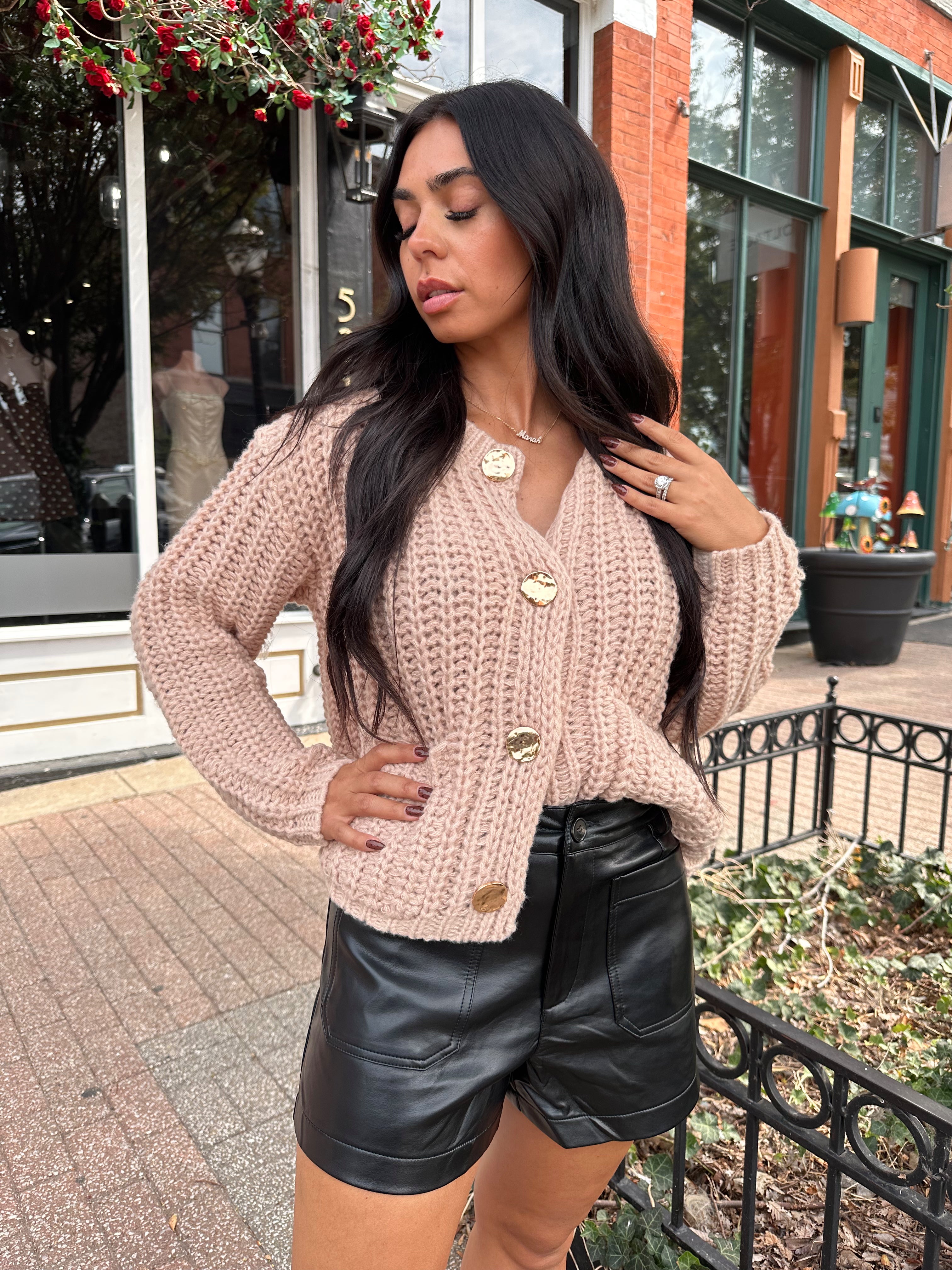 Woman wearing oatmeal colored knit chunky cardigan with gold button closure and functional pockets.