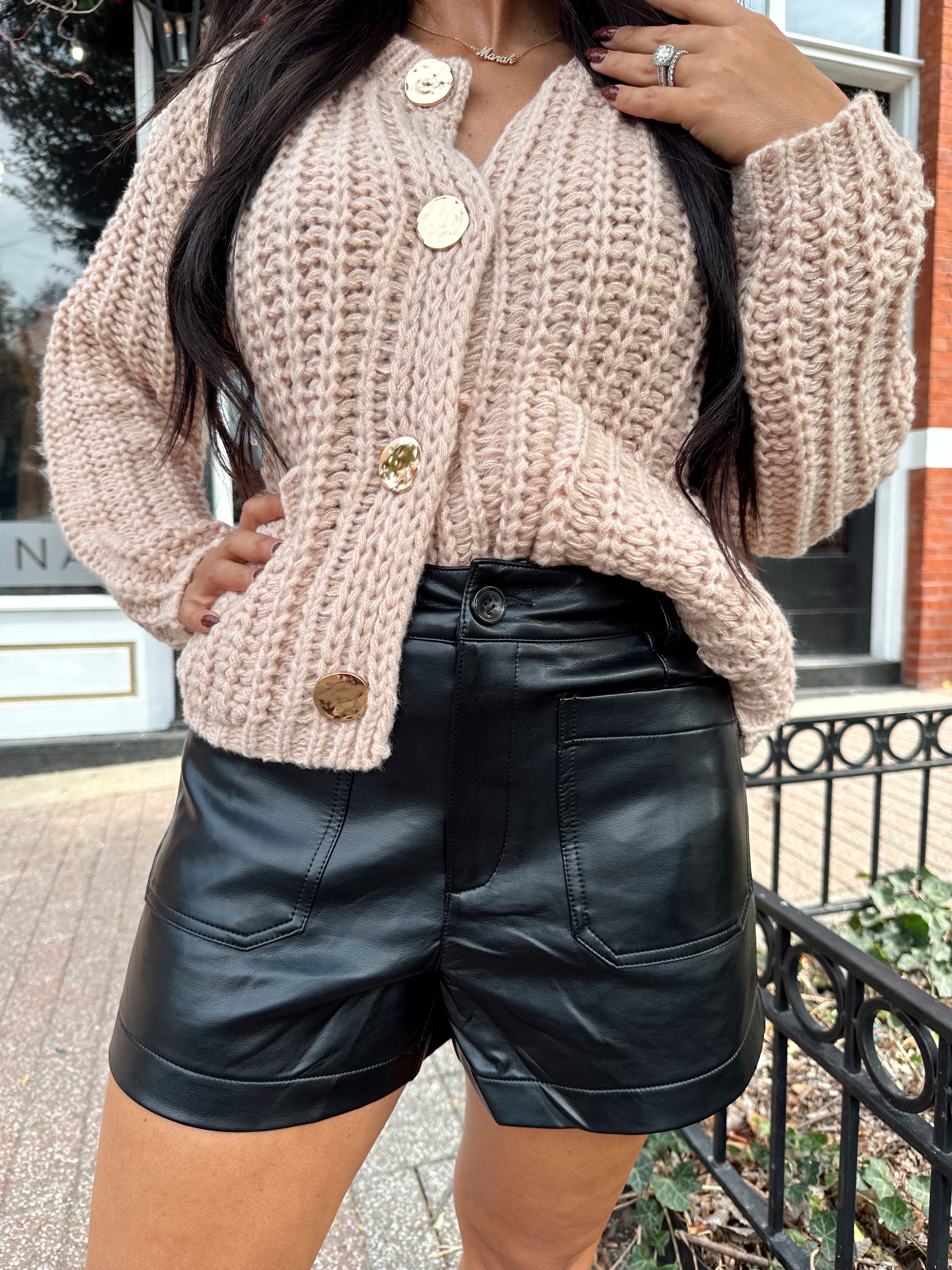 Woman wearing oatmeal colored knit chunky cardigan with gold button closure and functional pockets.