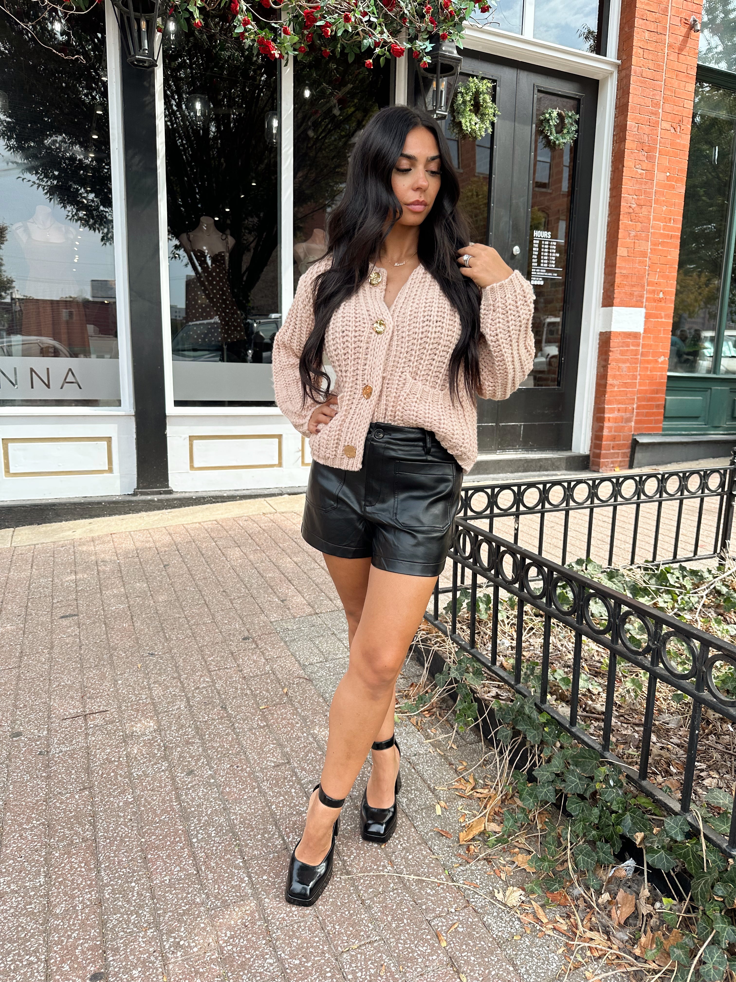 Woman wearing oatmeal colored knit chunky cardigan with gold button closure and functional pockets.