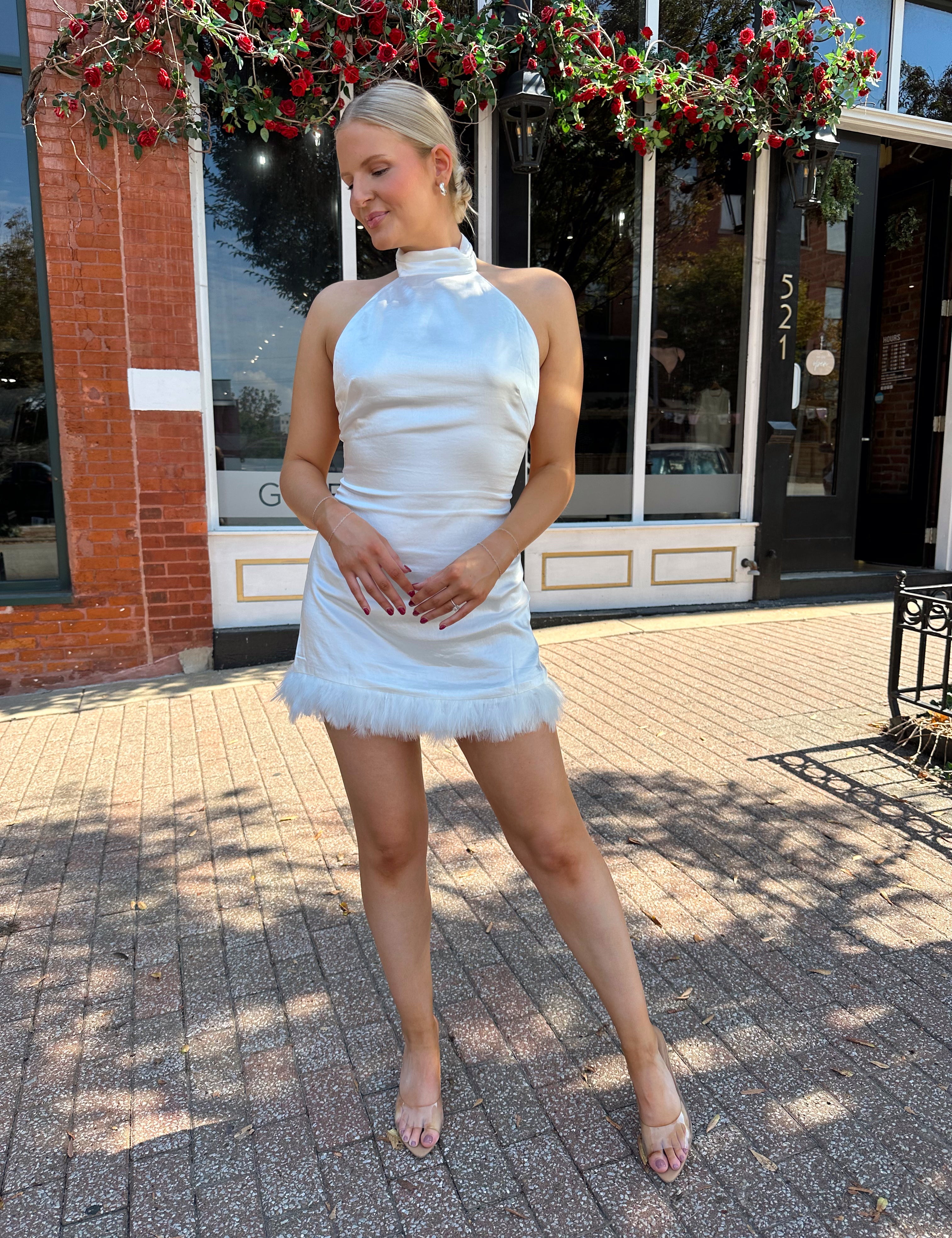 Woman wearing white high neck satin mini dress with open back, and feather trim.