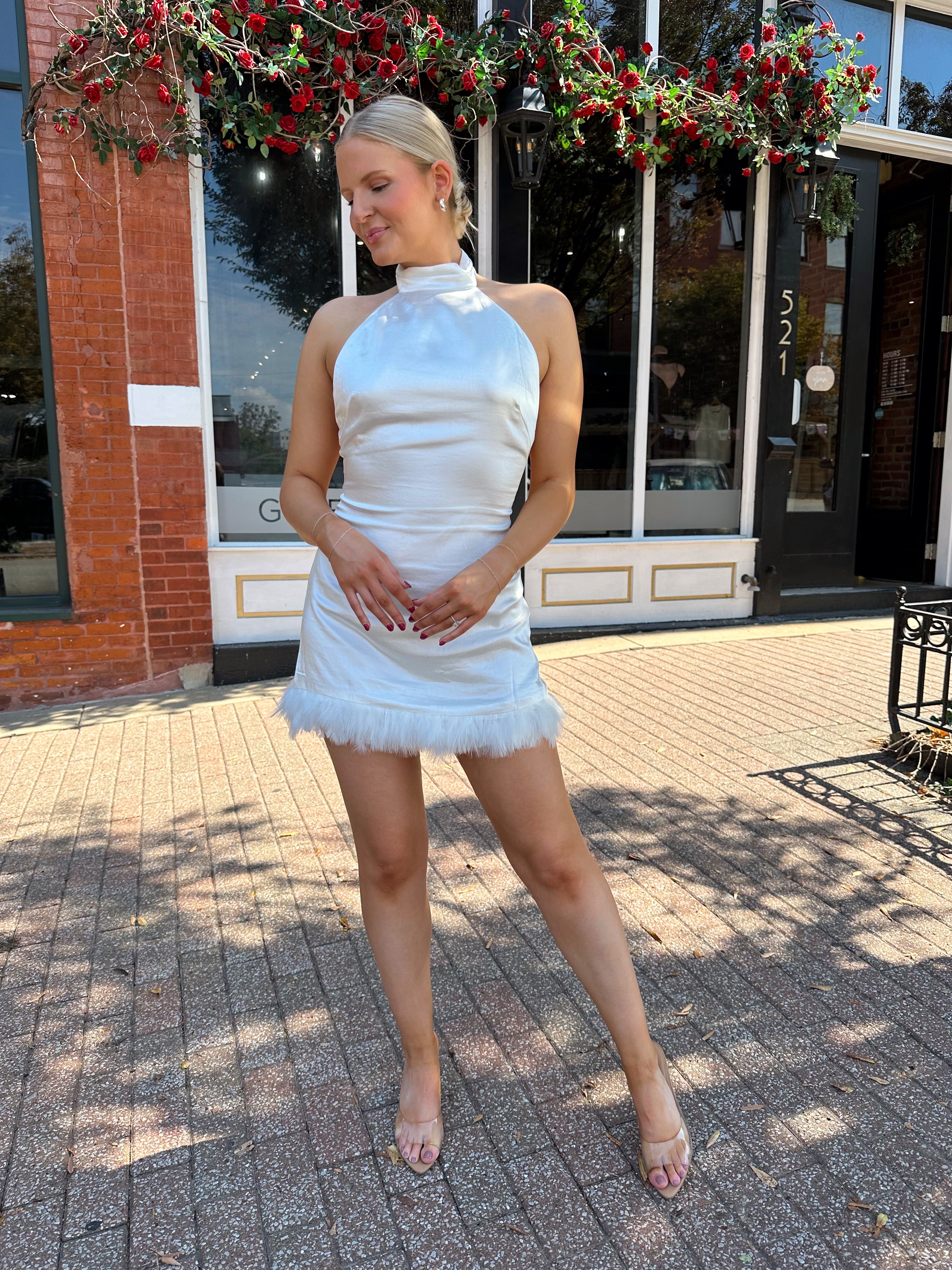 Woman wearing white high neck satin mini dress with open back, and feather trim.