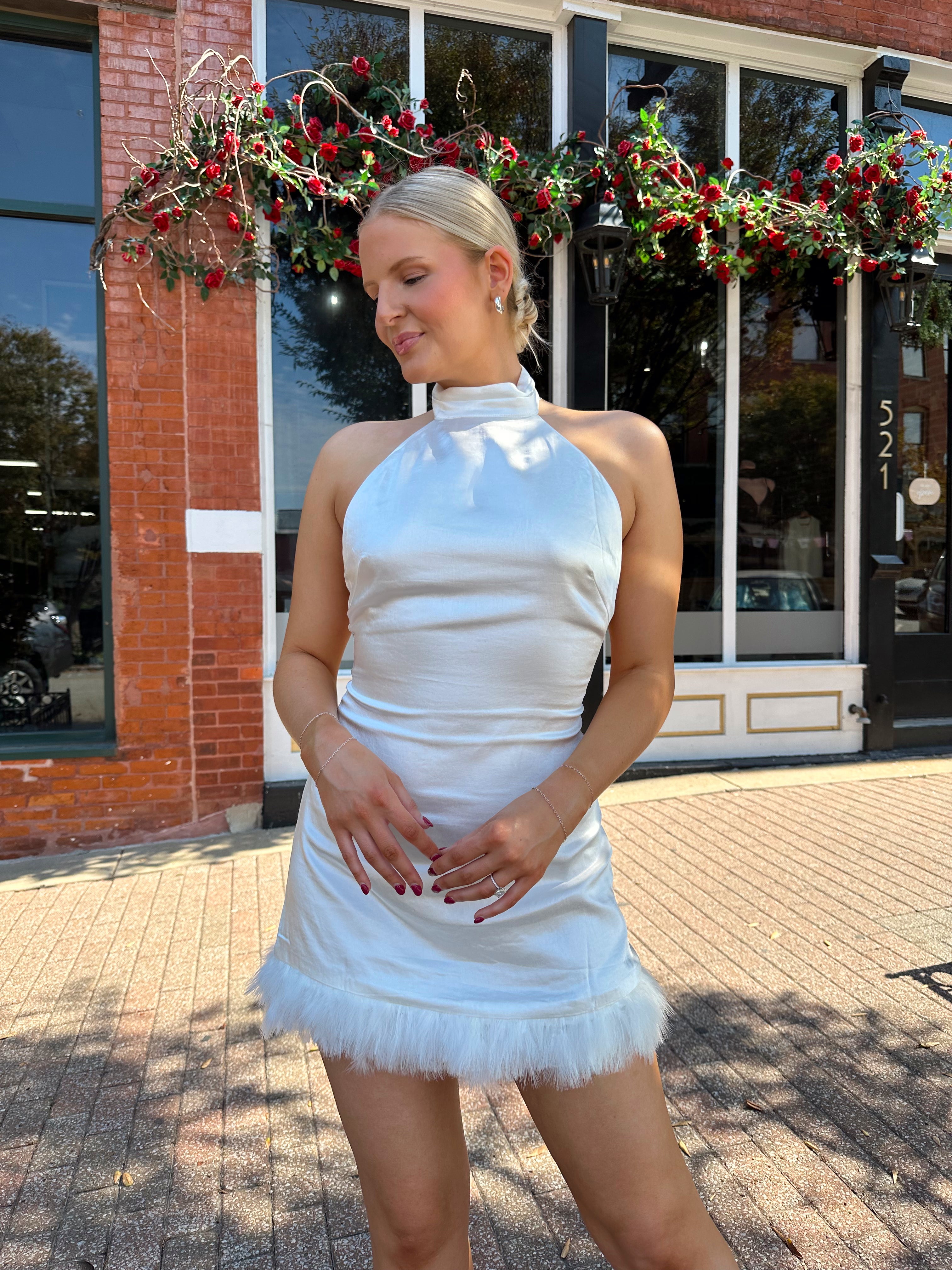 Woman wearing white high neck satin mini dress with open back, and feather trim.
