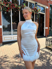Woman wearing white high neck satin mini dress with open back, and feather trim.