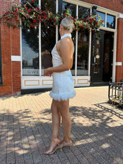 Woman wearing white high neck satin mini dress with open back, and feather trim.