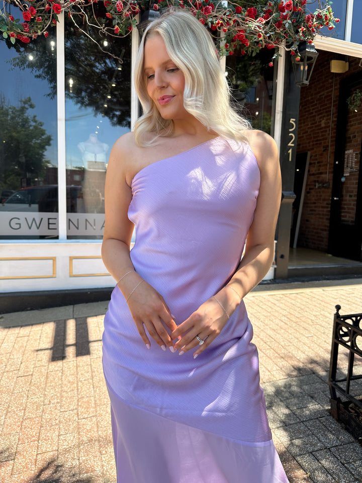 Woman wearing one shoulder lavender maxi dress with sheer leg paneling, open back, and strap detailing.