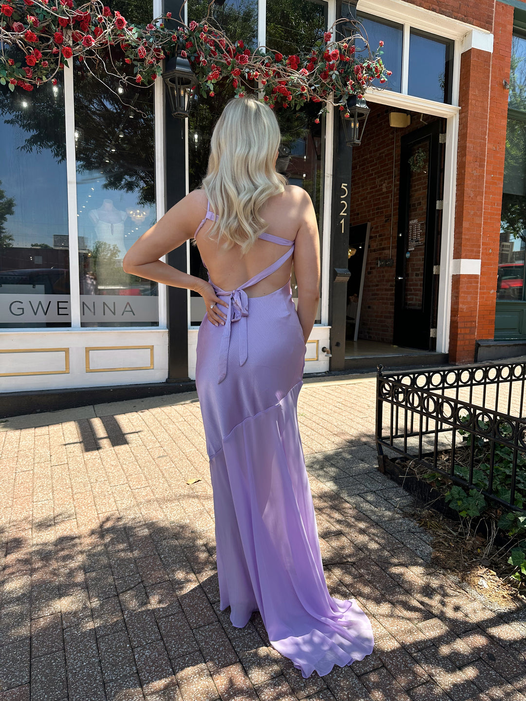 Woman wearing one shoulder lavender maxi dress with sheer leg paneling, open back, and strap detailing.