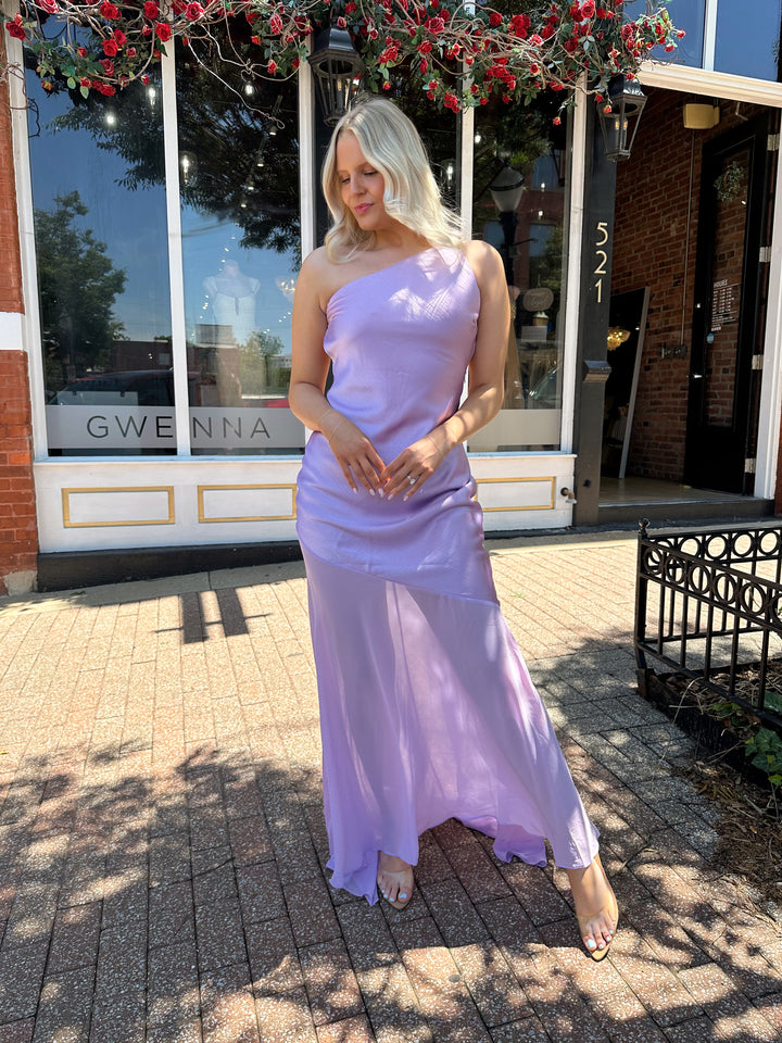 Woman wearing one shoulder lavender maxi dress with sheer leg paneling, open back, and strap detailing.