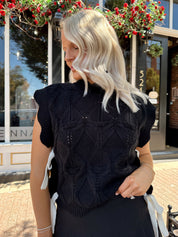 Woman wearing black cable knit sleeveless sweater with contrast bow back. 