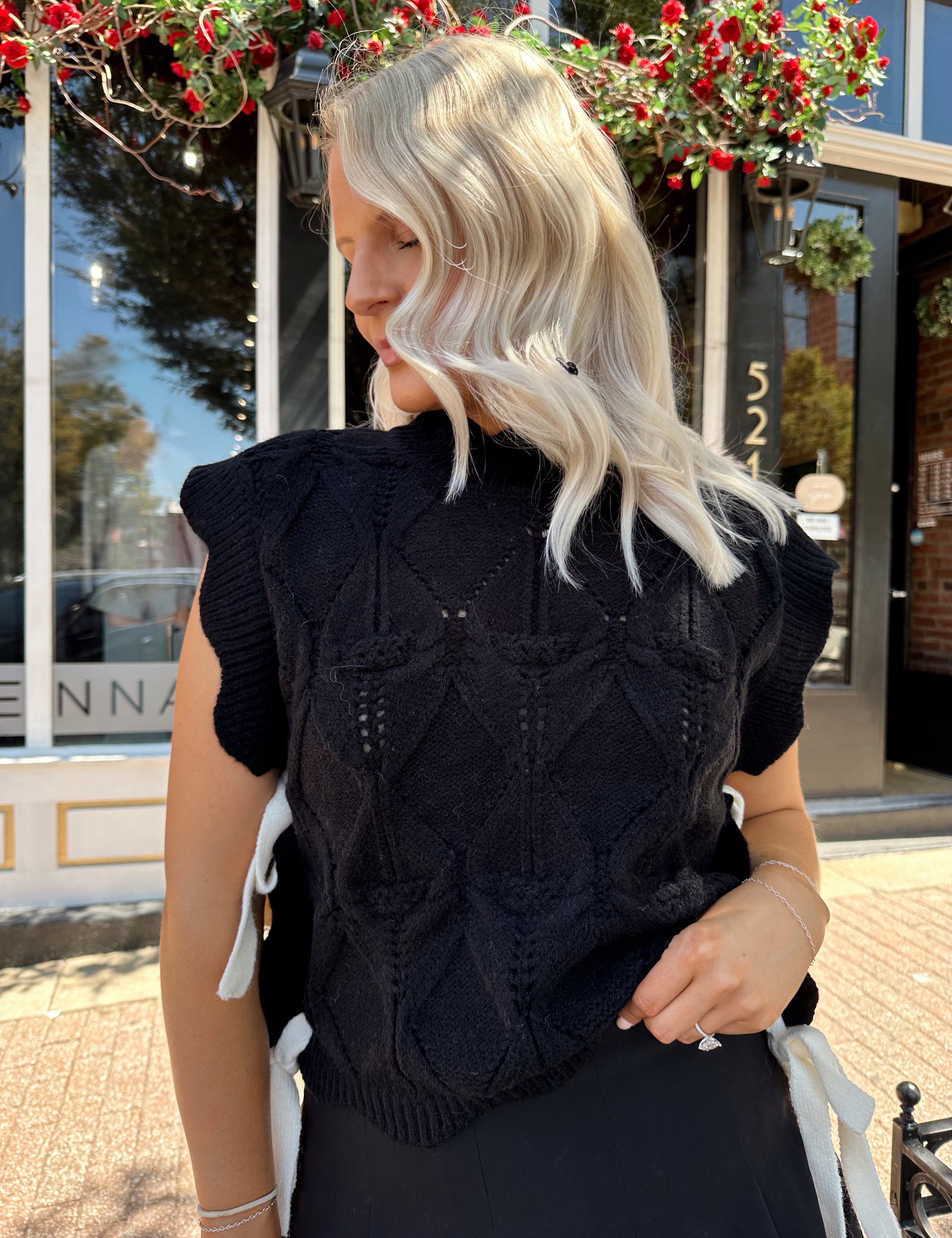 Woman wearing black cable knit sleeveless sweater with contrast bow back. 