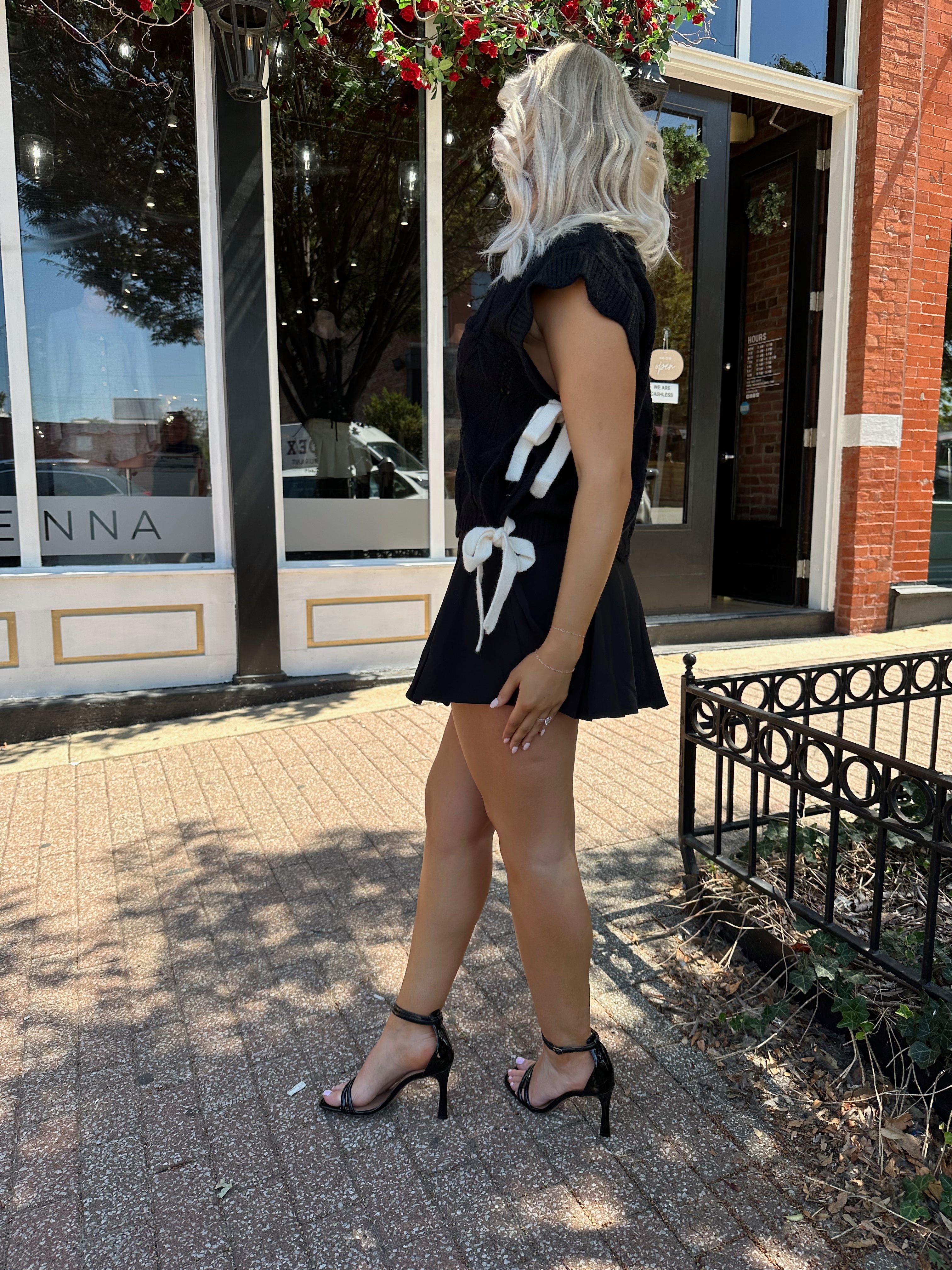 Woman wearing black cable knit sleeveless sweater with contrast bow back. 
