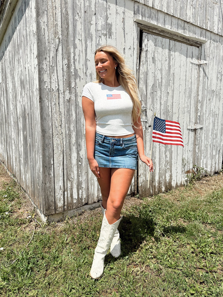 Woman wearing a light grey baby tee with an embroidered American flag on the chest