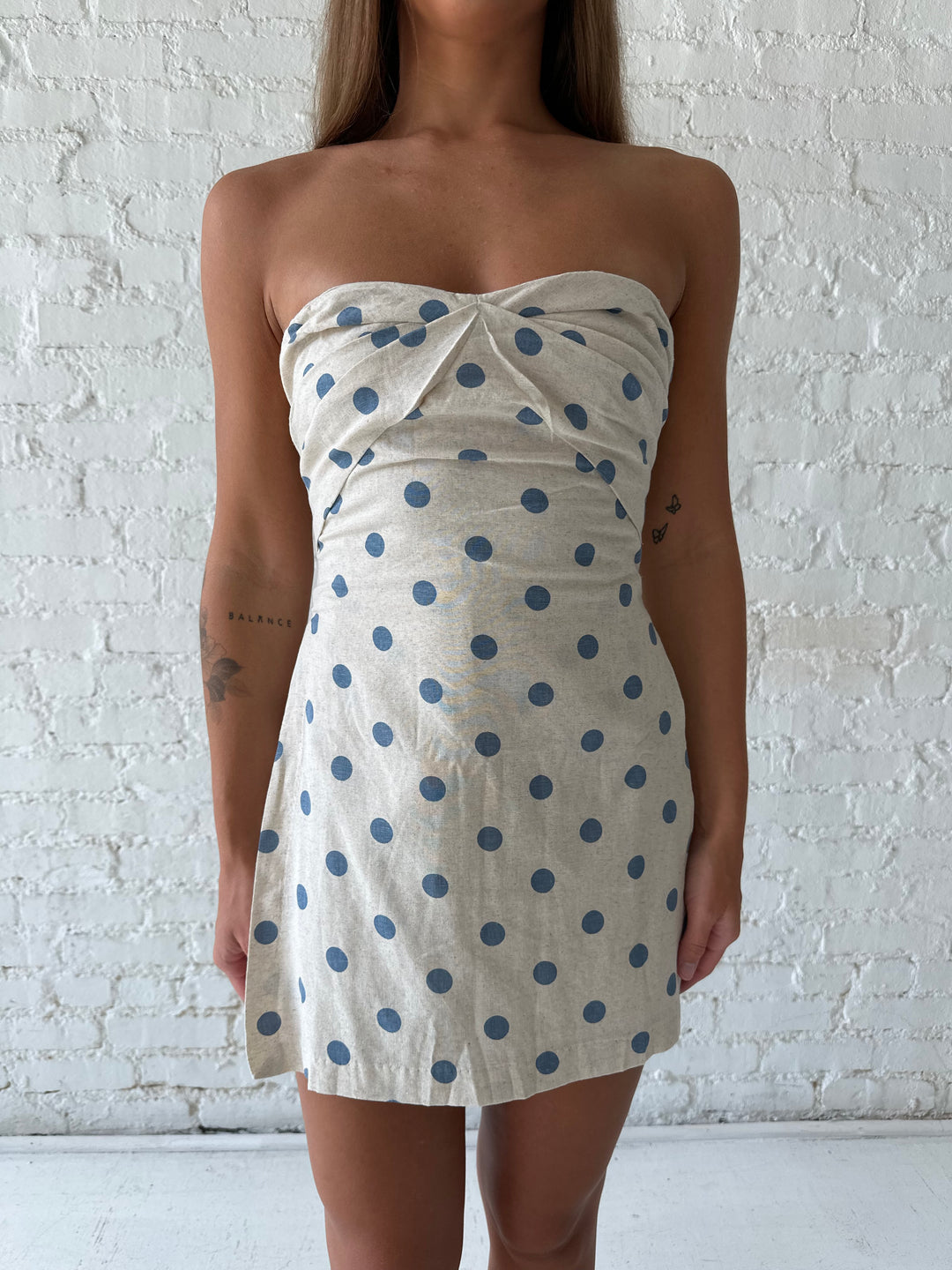Woman wearing a strapless oatmeal linen romper with blue polka dots, and side slits. 
