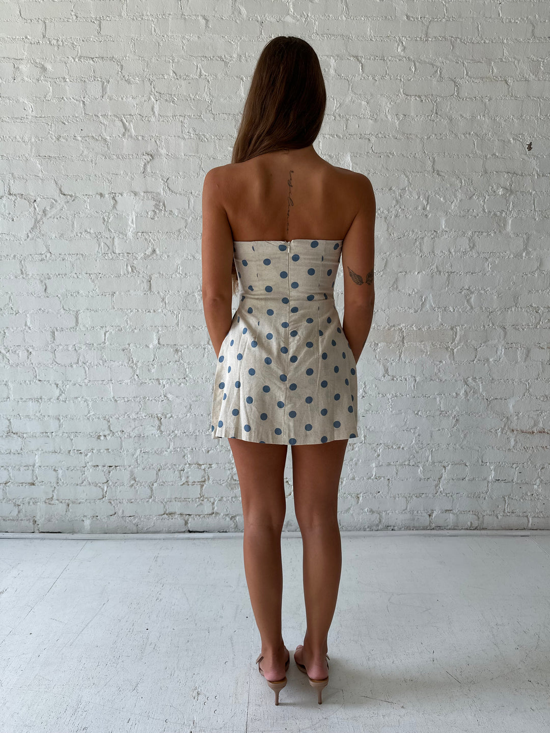 Woman wearing a strapless oatmeal linen romper with blue polka dots, and side slits. 