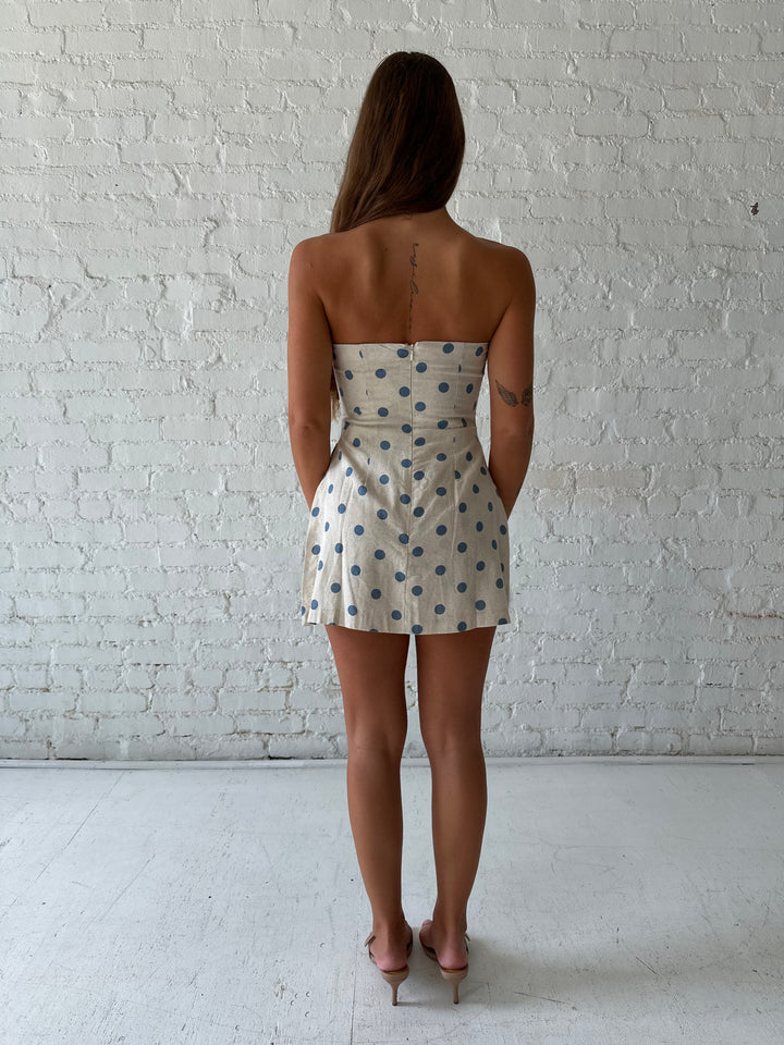 Woman wearing a strapless oatmeal linen romper with blue polka dots, and side slits. 