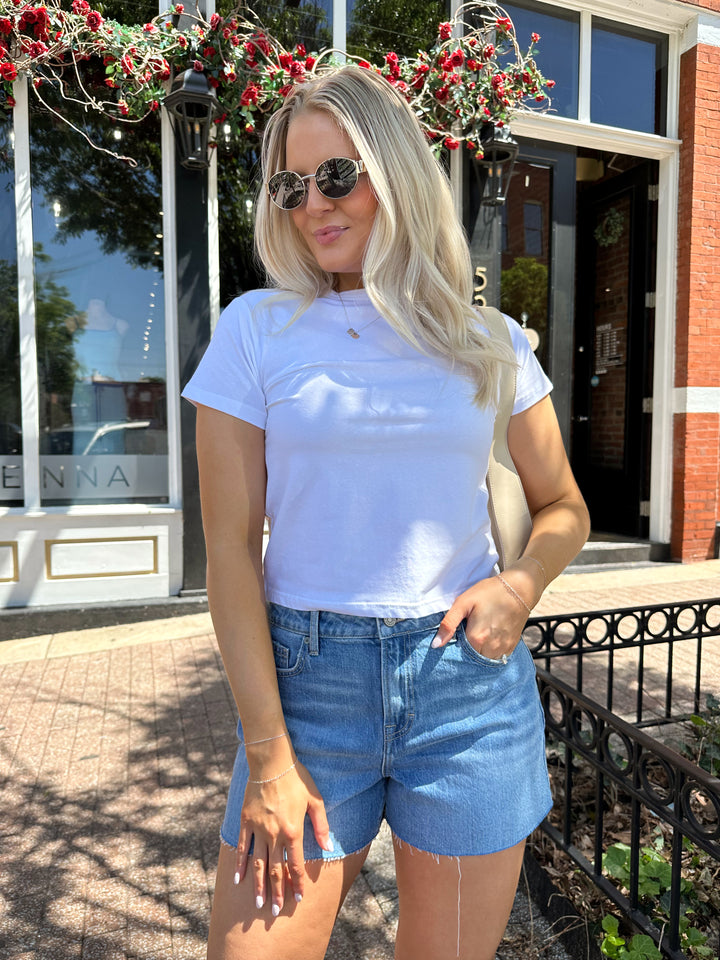 Woman wearing medium wash denim shorts with raw hem, and white t-shirt. 