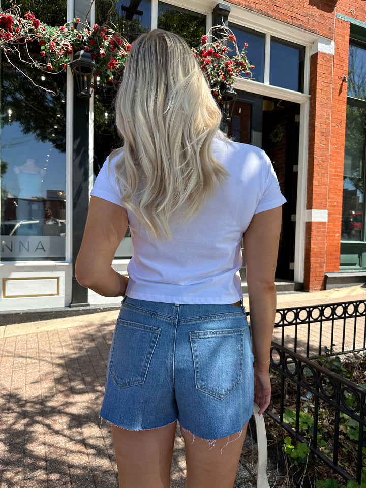 Woman wearing medium wash denim shorts with raw hem, and white t-shirt. 