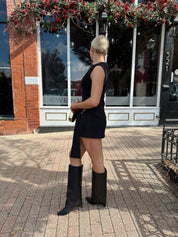 Woman wearing black mini dress with gold emblem and belt sash with black knee boot.