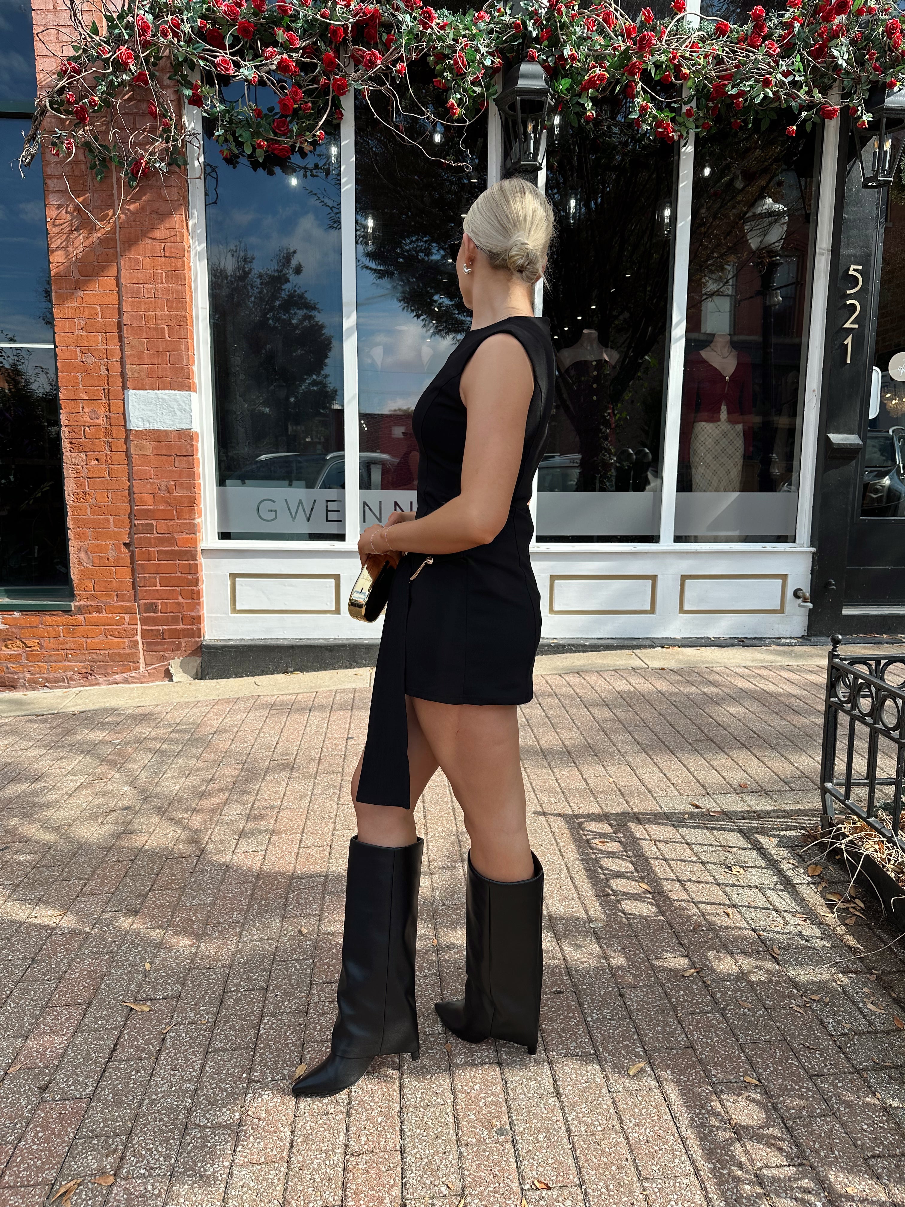 Woman wearing black mini dress with gold emblem and belt sash with black knee boot.