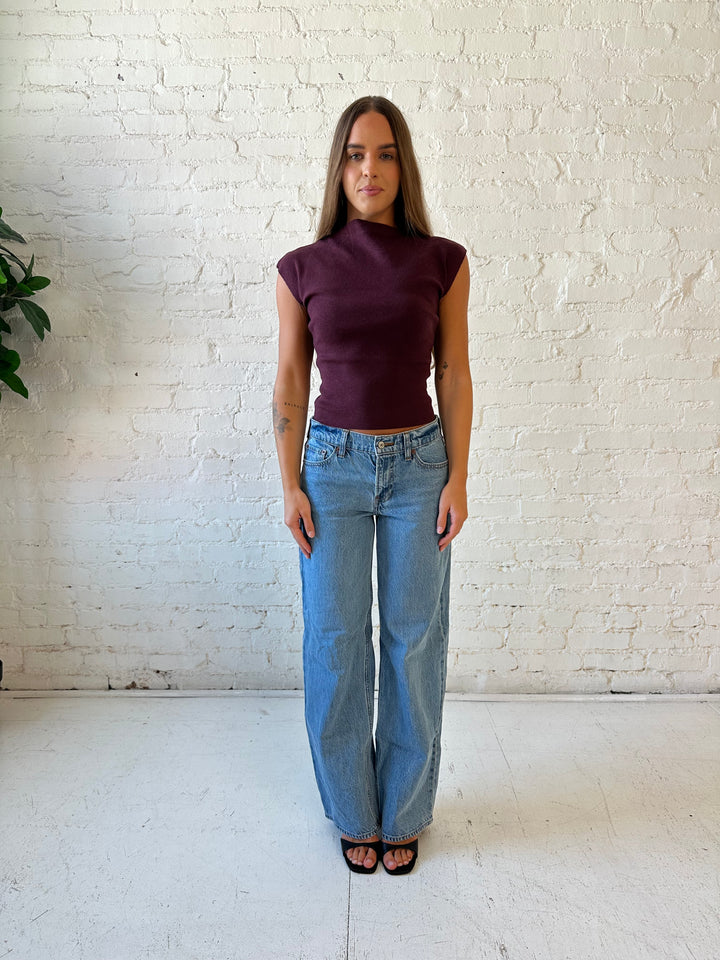Woman wearing plum colored ribbed cap sleeve top with high neckline, and medium wash jean. 