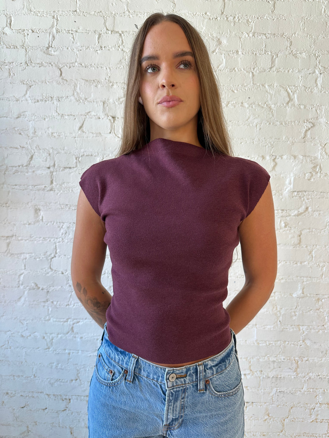 Woman wearing plum colored ribbed cap sleeve top with high neckline, and medium wash jean. 