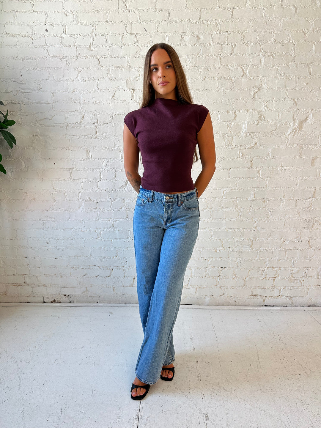 Woman wearing plum colored ribbed cap sleeve top with high neckline, and medium wash jean. 