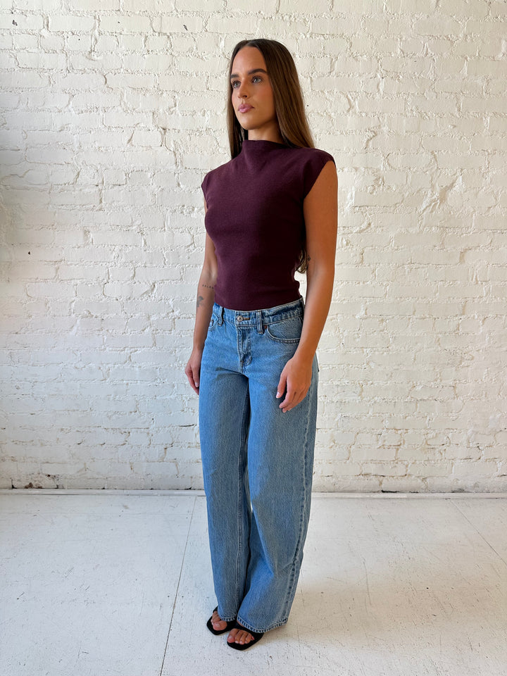 Woman wearing plum colored ribbed cap sleeve top with high neckline, and medium wash jean. 