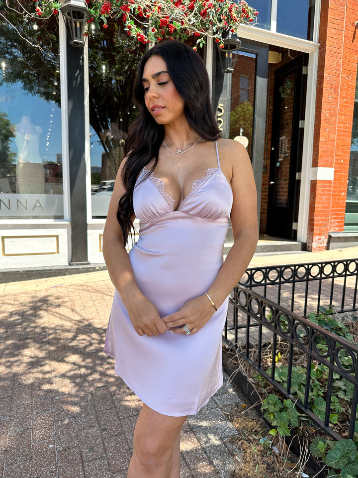 Woman wearing lavender satin mini dress with lace trim detailing, open back, and self tie criss-cross straps.
