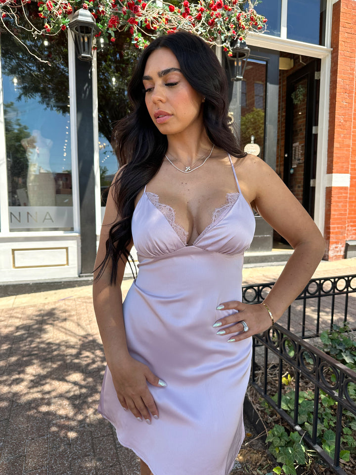 Woman wearing lavender satin mini dress with lace trim detailing, open back, and self tie criss-cross straps.