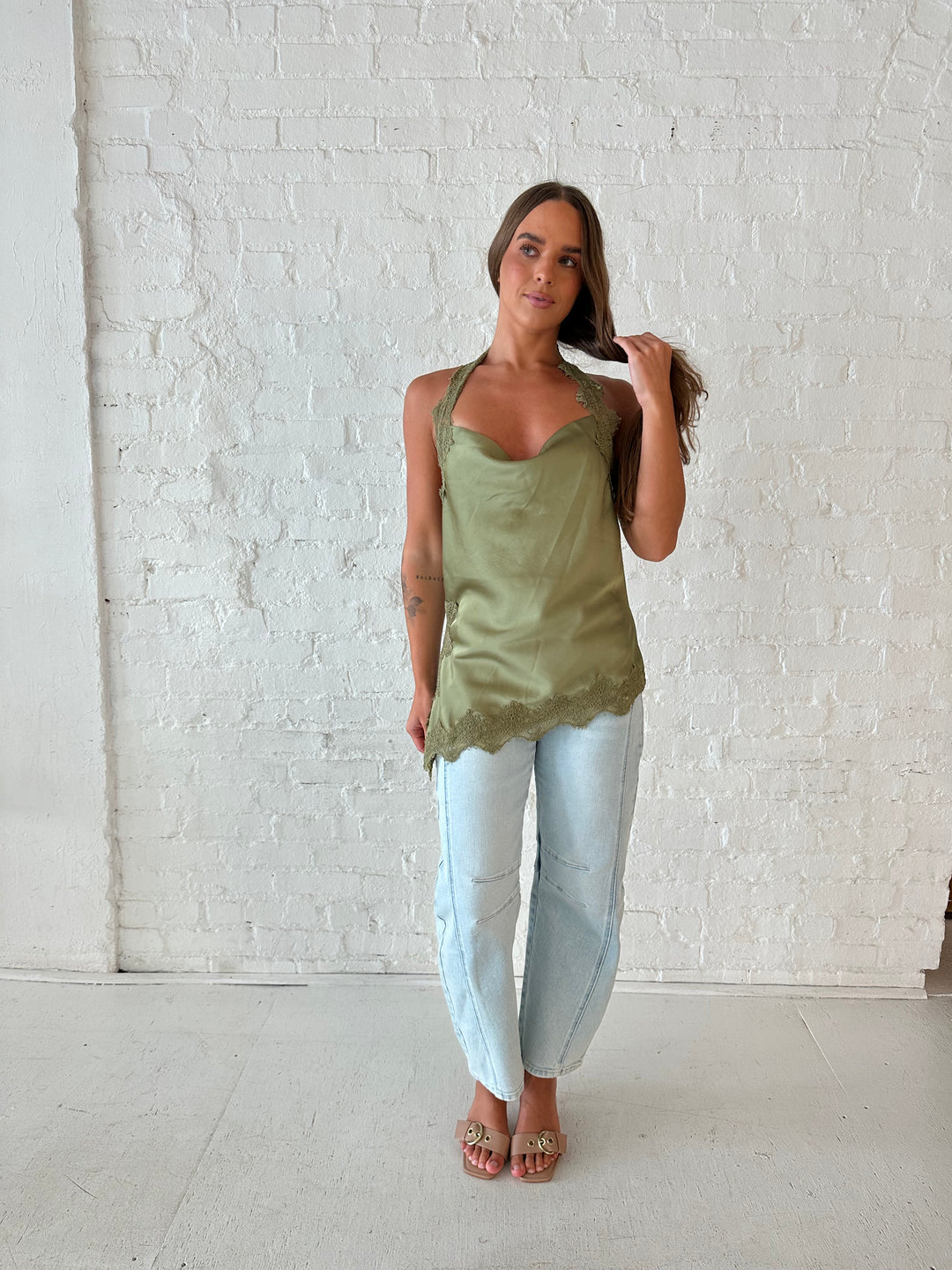 Woman wearing sage green satin lace trim top with asymmetrical hem, and open back. 
