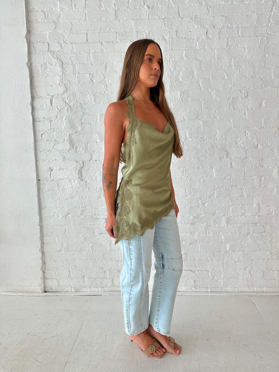Woman wearing sage green satin lace trim top with asymmetrical hem, and open back. 