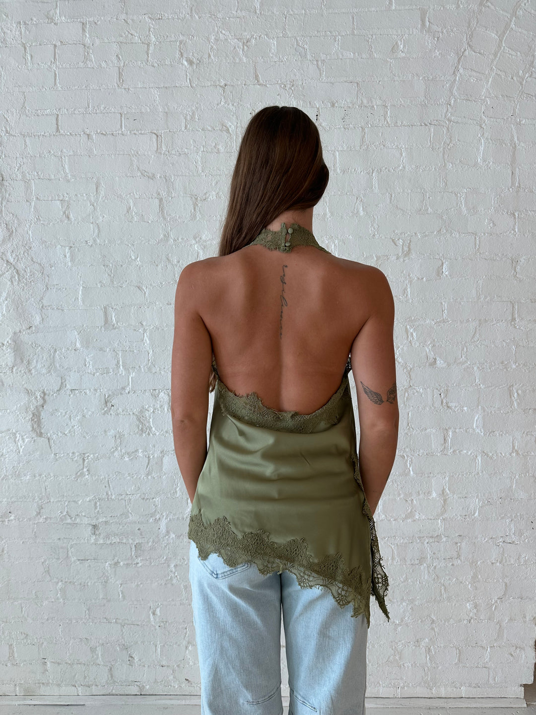 Woman wearing sage green satin lace trim top with asymmetrical hem, and open back. 