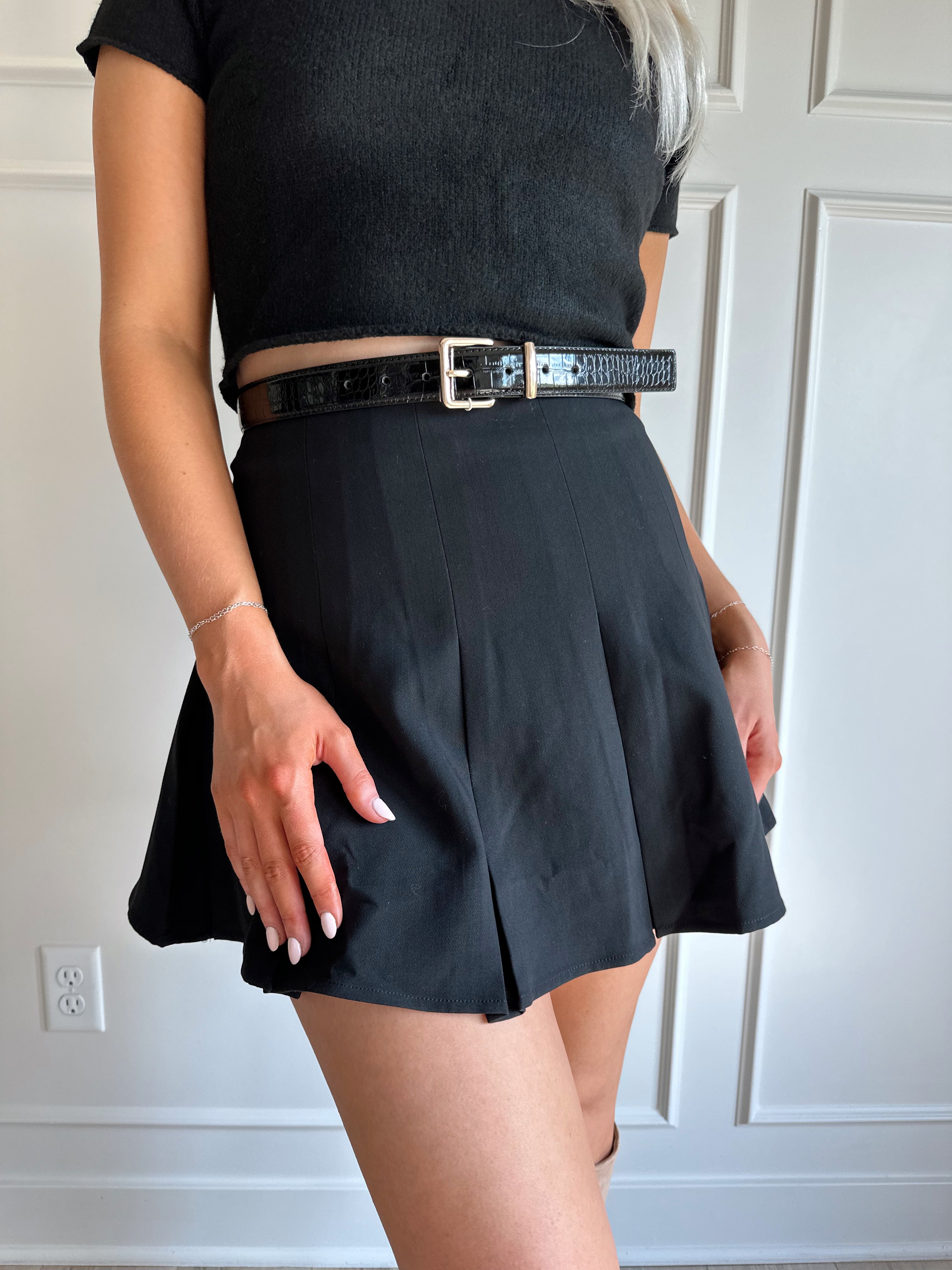 Woman wearing a black pleated mini skirt with black top and black croc belt.