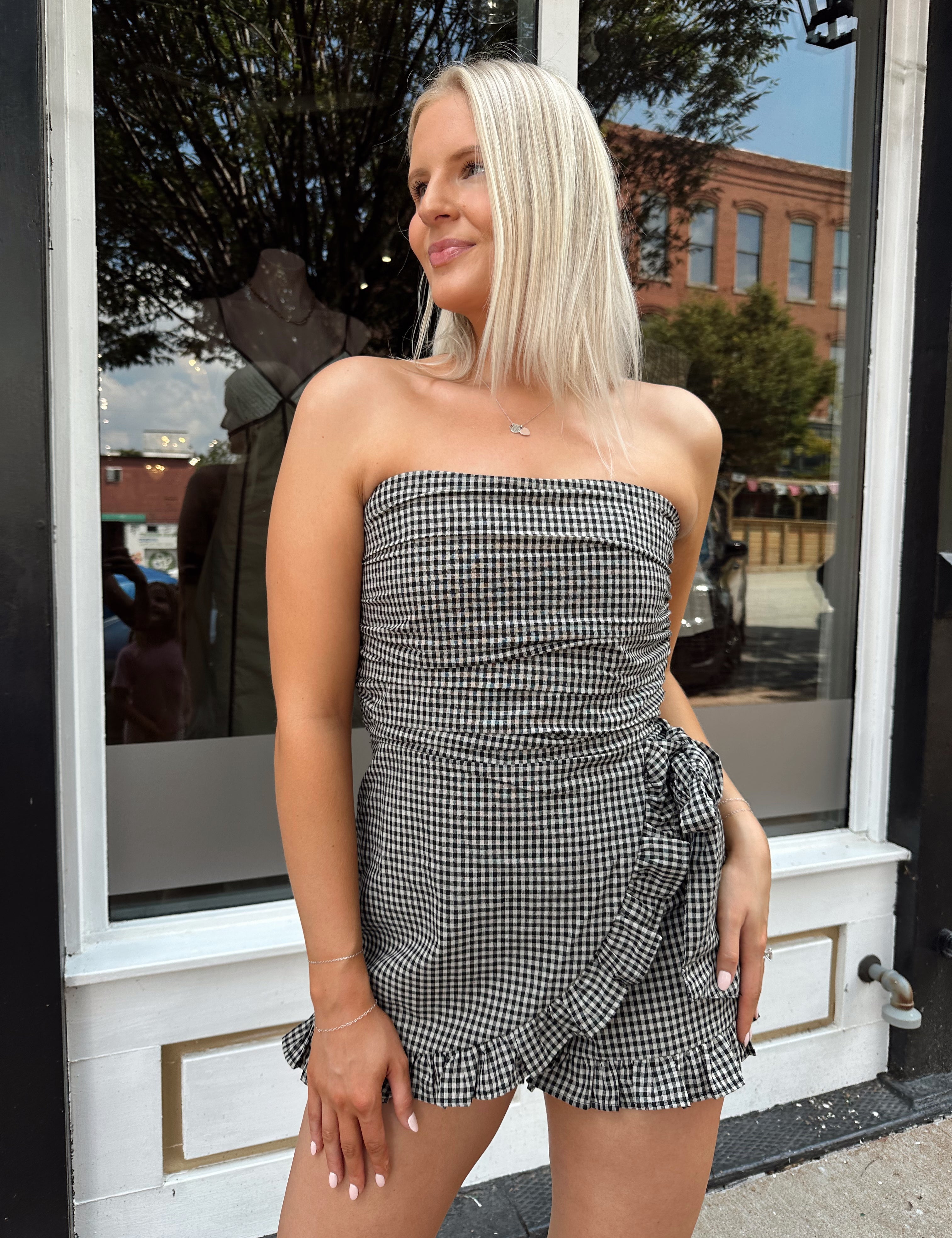 Woman wearing a black and taupe gingham strapless romper with wrap skirt. 