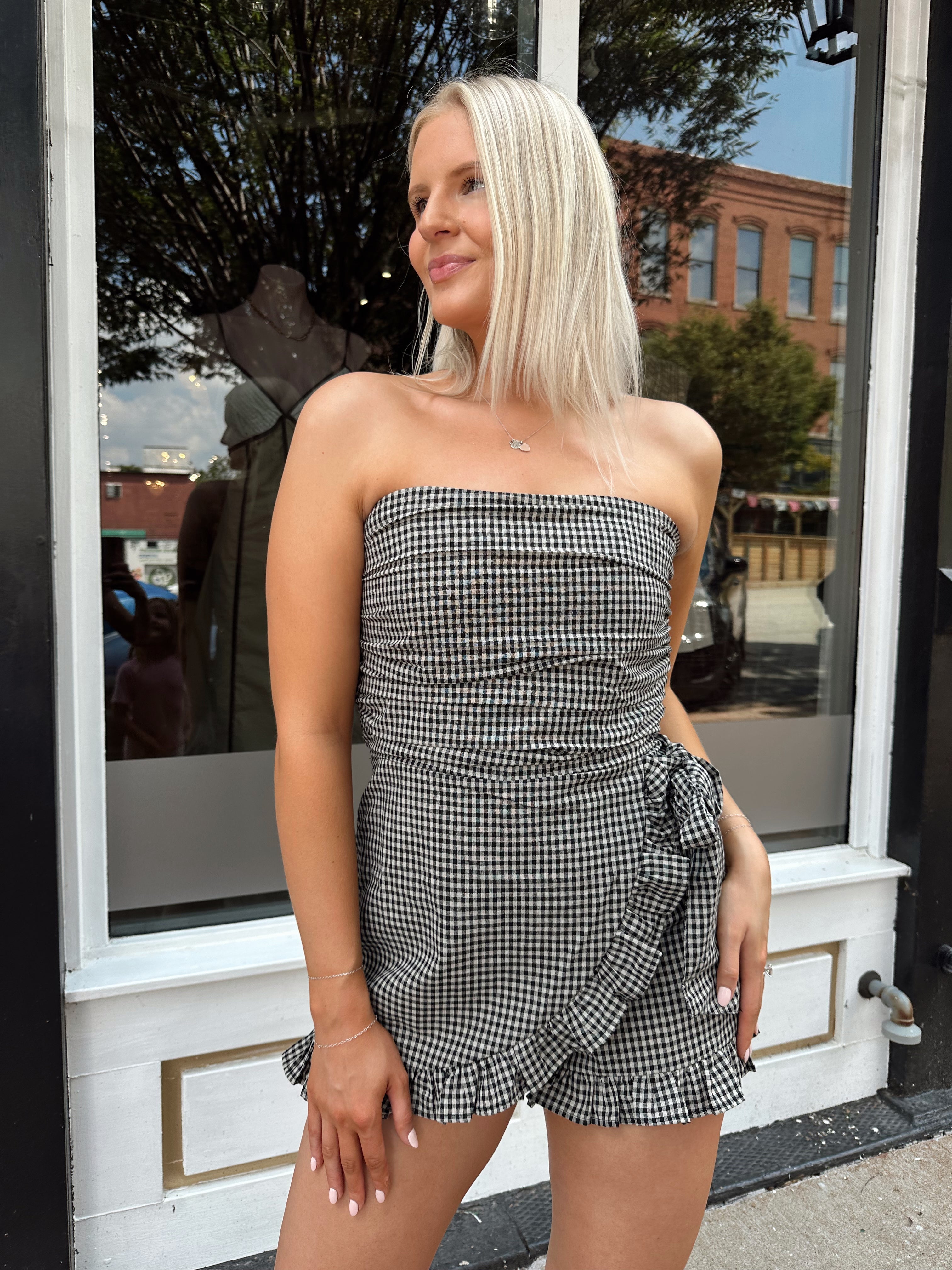 Woman wearing a black and taupe gingham strapless romper with wrap skirt. 