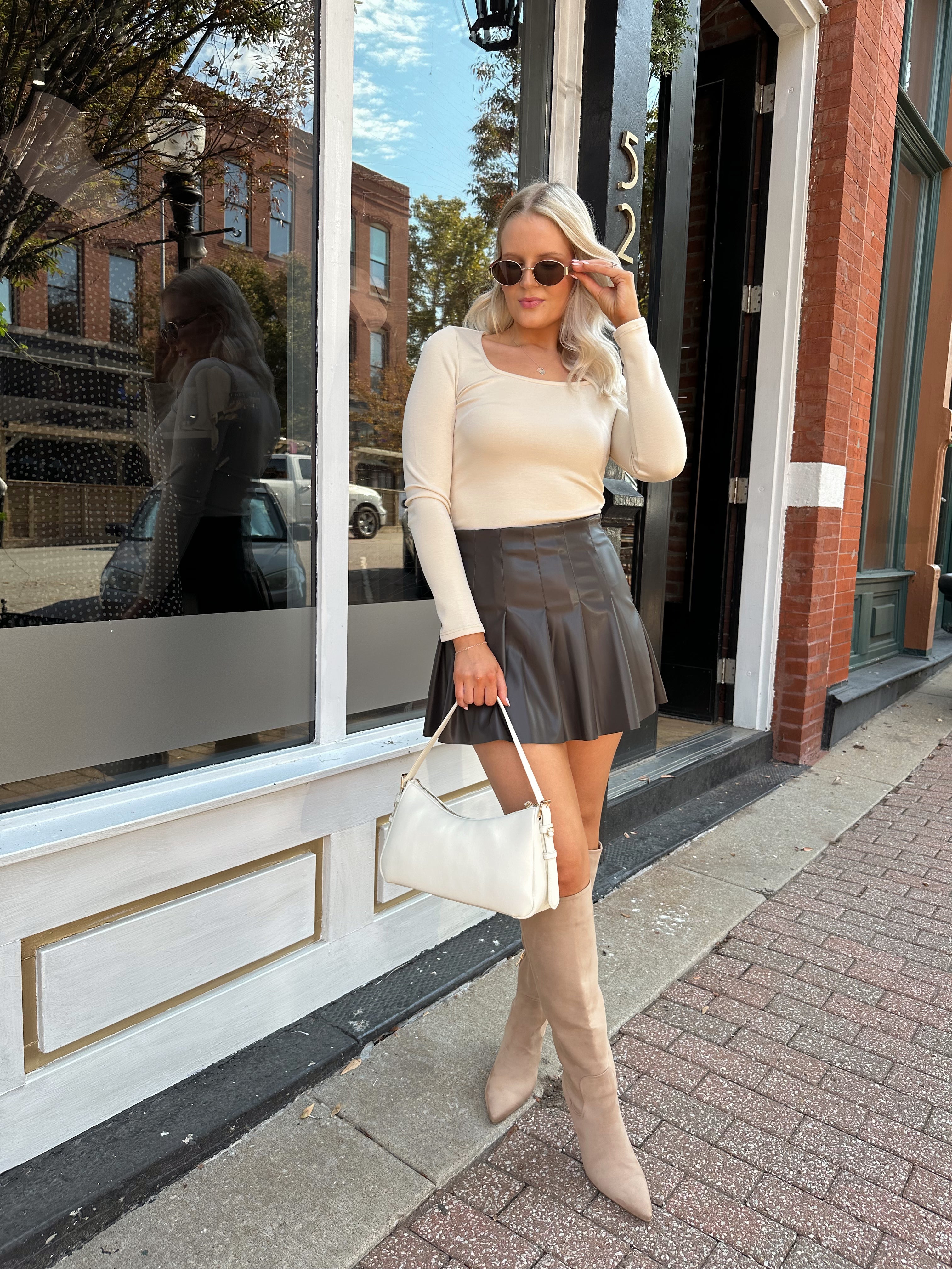 Woman wearing cream long sleeve scoop neck top with brown pleated mini skirt.