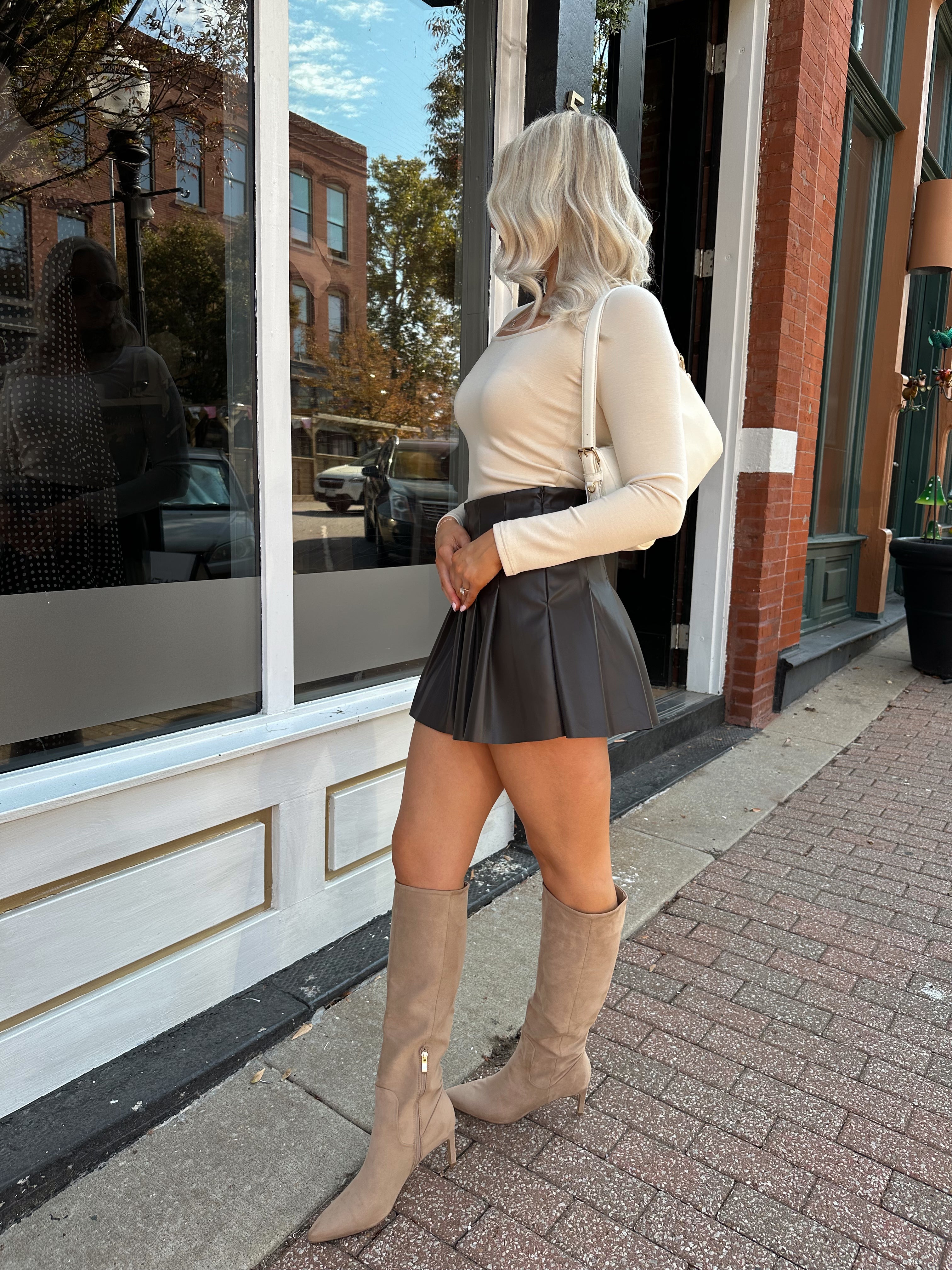 Woman wearing cream long sleeve scoop neck top with brown pleated mini skirt.