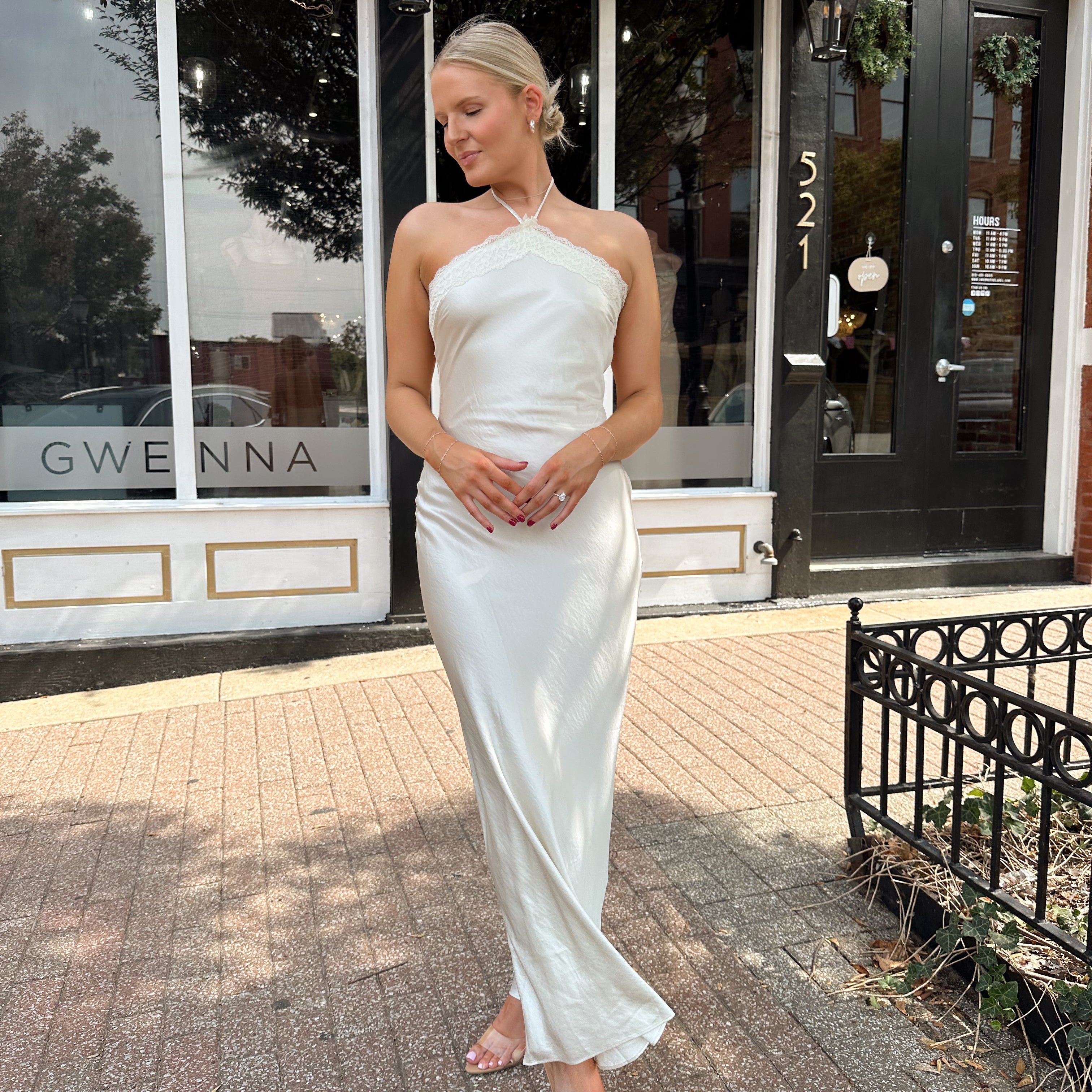 Woman wearing off white satin maxi dress with lace trim detailing, and self tie halter neck. 