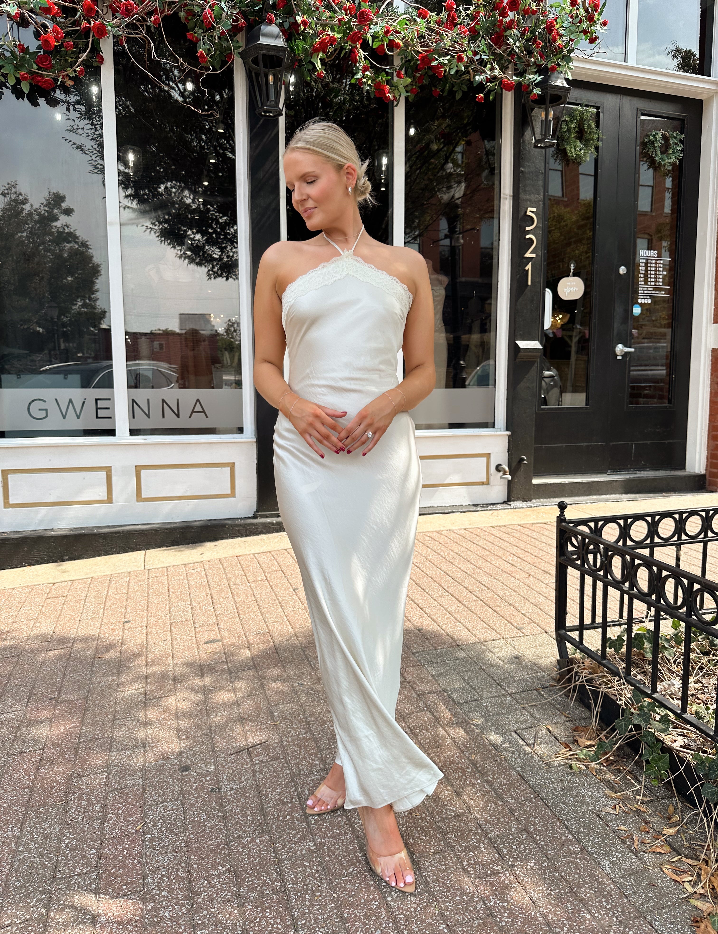 Woman wearing off white satin maxi dress with lace trim detailing, and self tie halter neck. 