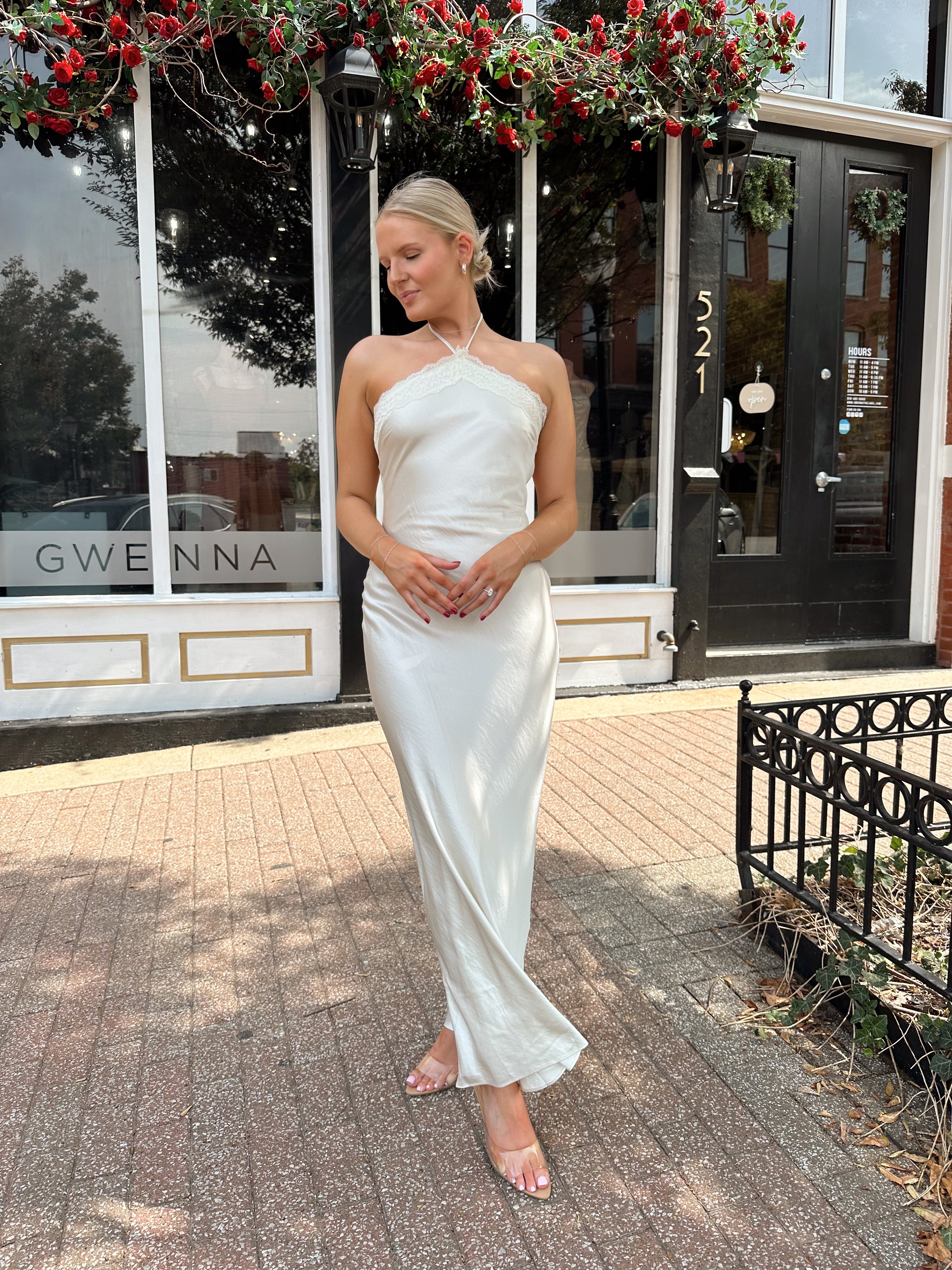 Woman wearing off white satin maxi dress with lace trim detailing, and self tie halter neck. 