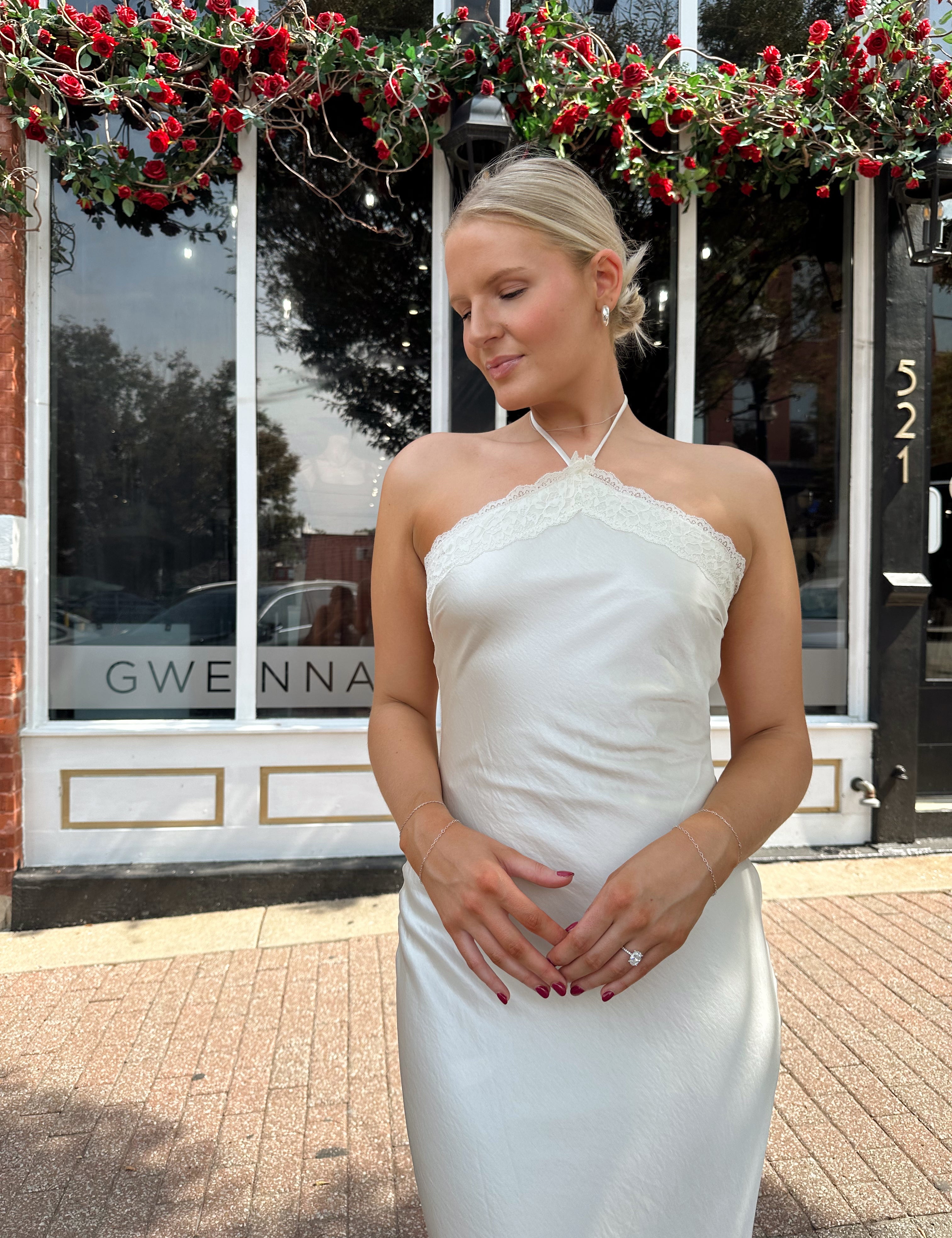 Woman wearing off white satin maxi dress with lace trim detailing, and self tie halter neck. 