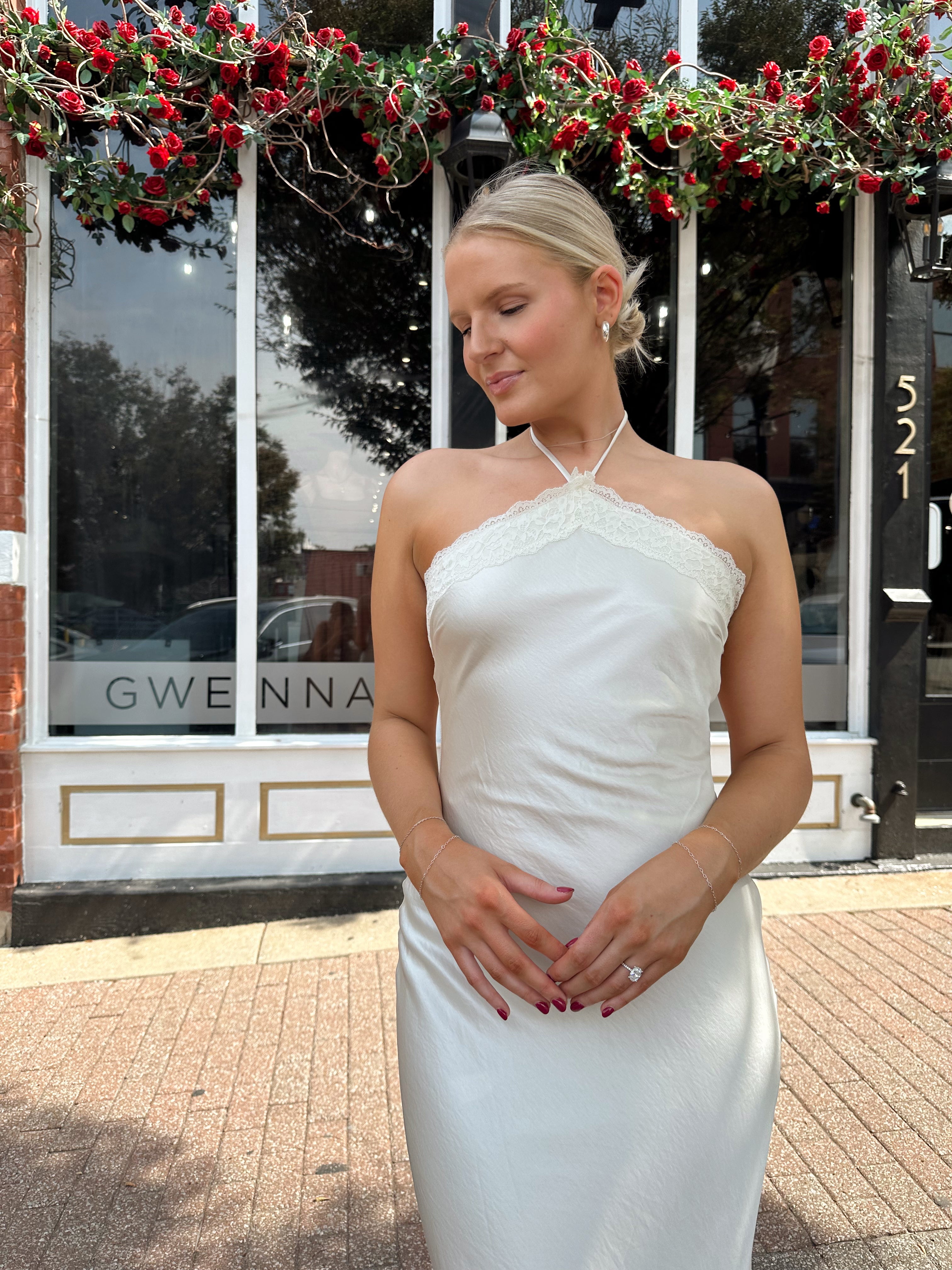 Woman wearing off white satin maxi dress with lace trim detailing, and self tie halter neck. 