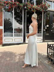 Woman wearing off white satin maxi dress with lace trim detailing, and self tie halter neck. 