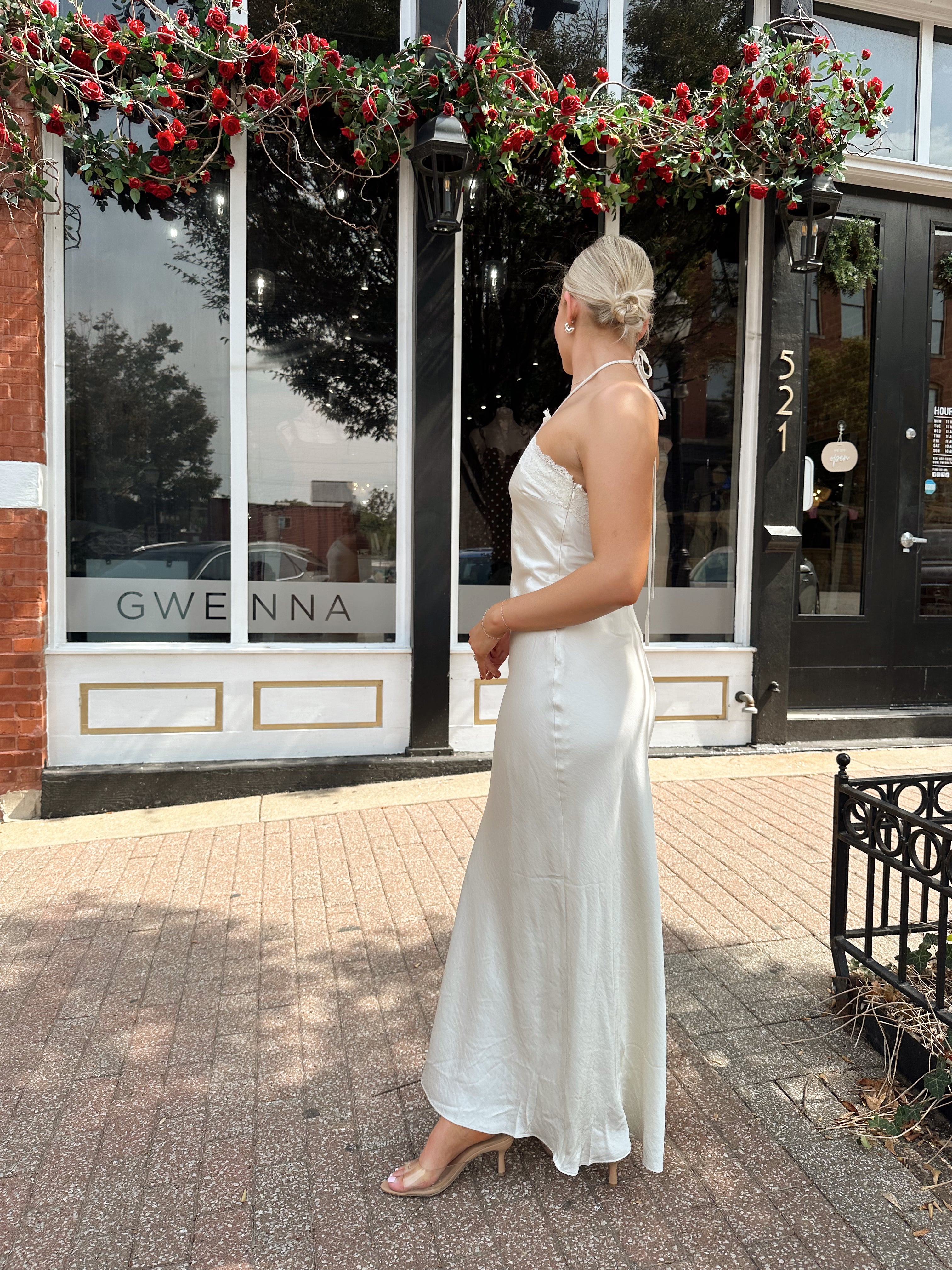 Woman wearing off white satin maxi dress with lace trim detailing, and self tie halter neck. 