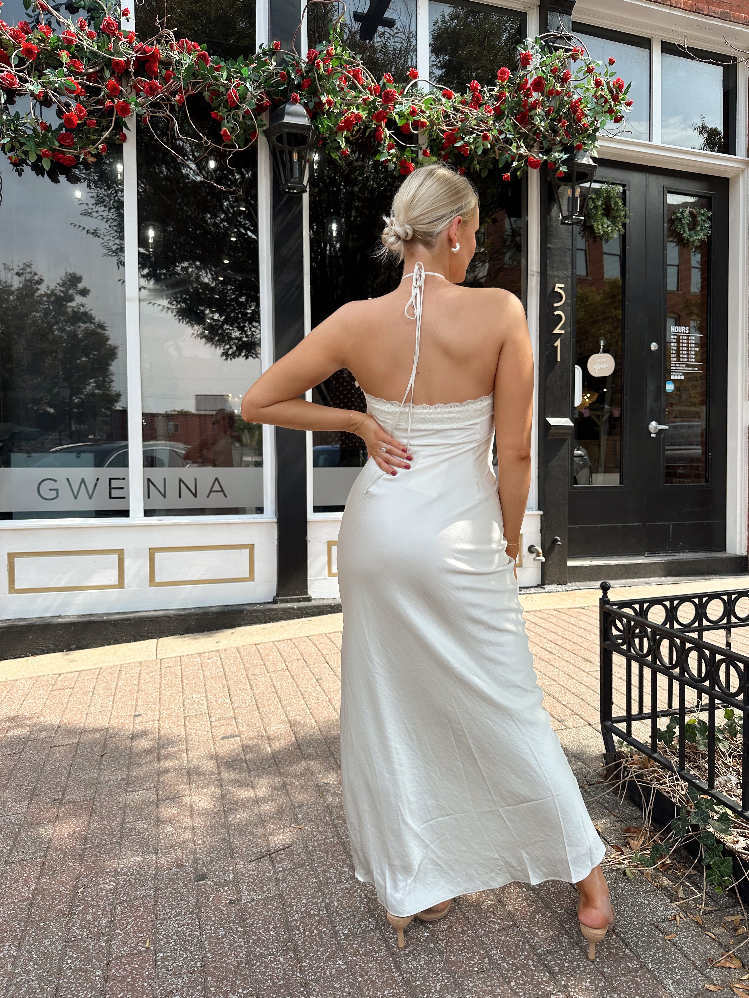 Woman wearing off white satin maxi dress with lace trim detailing, and self tie halter neck. 