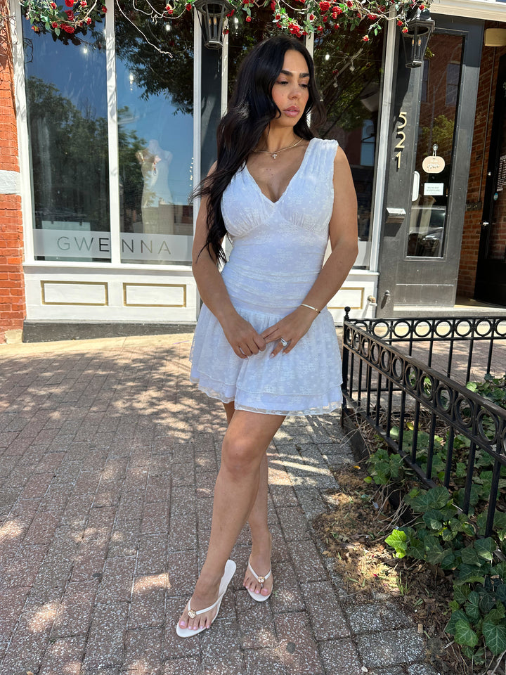 Woman wearing white lace mini dress with drop waist, v-neckline, and open back with tie detail. 
