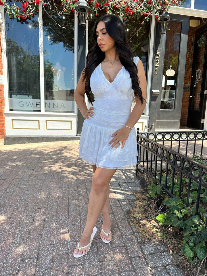Woman wearing white lace mini dress with drop waist, v-neckline, and open back with tie detail. 