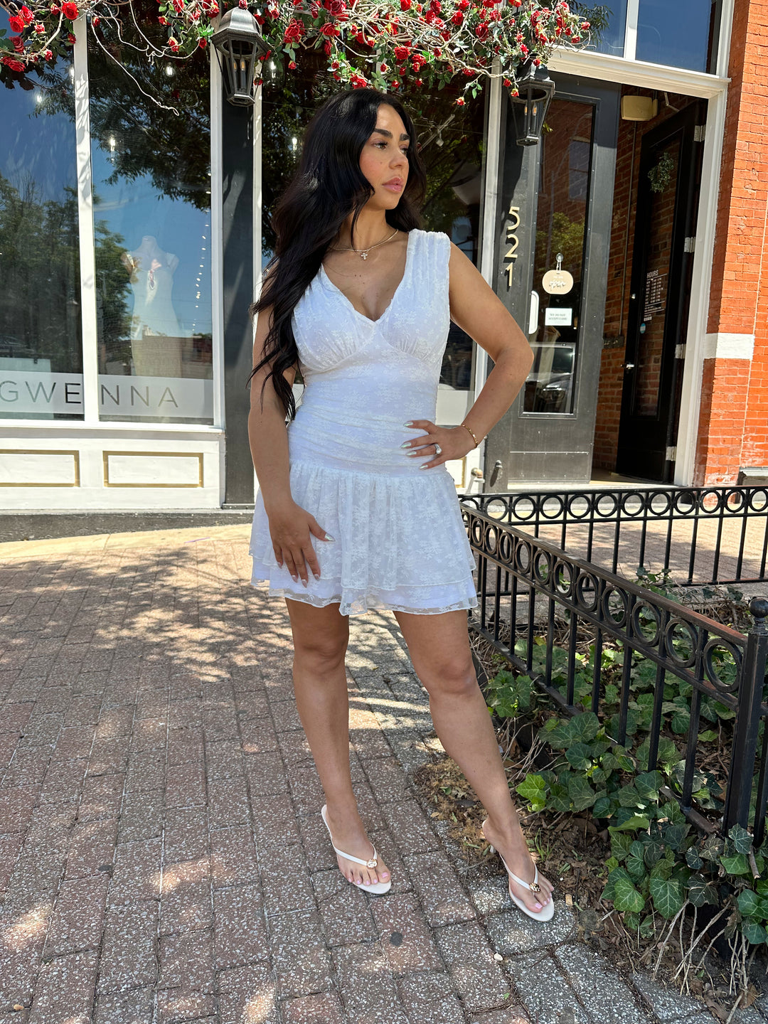 Woman wearing white lace mini dress with drop waist, v-neckline, and open back with tie detail. 