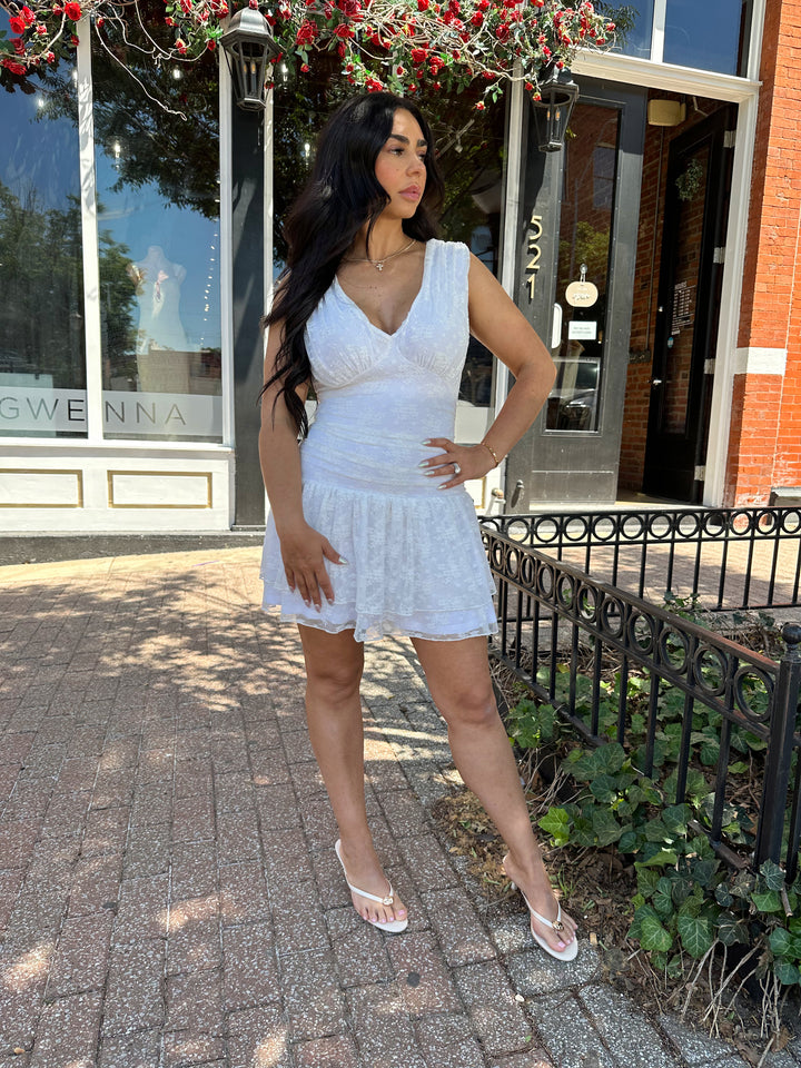 Woman wearing white lace mini dress with drop waist, v-neckline, and open back with tie detail. 